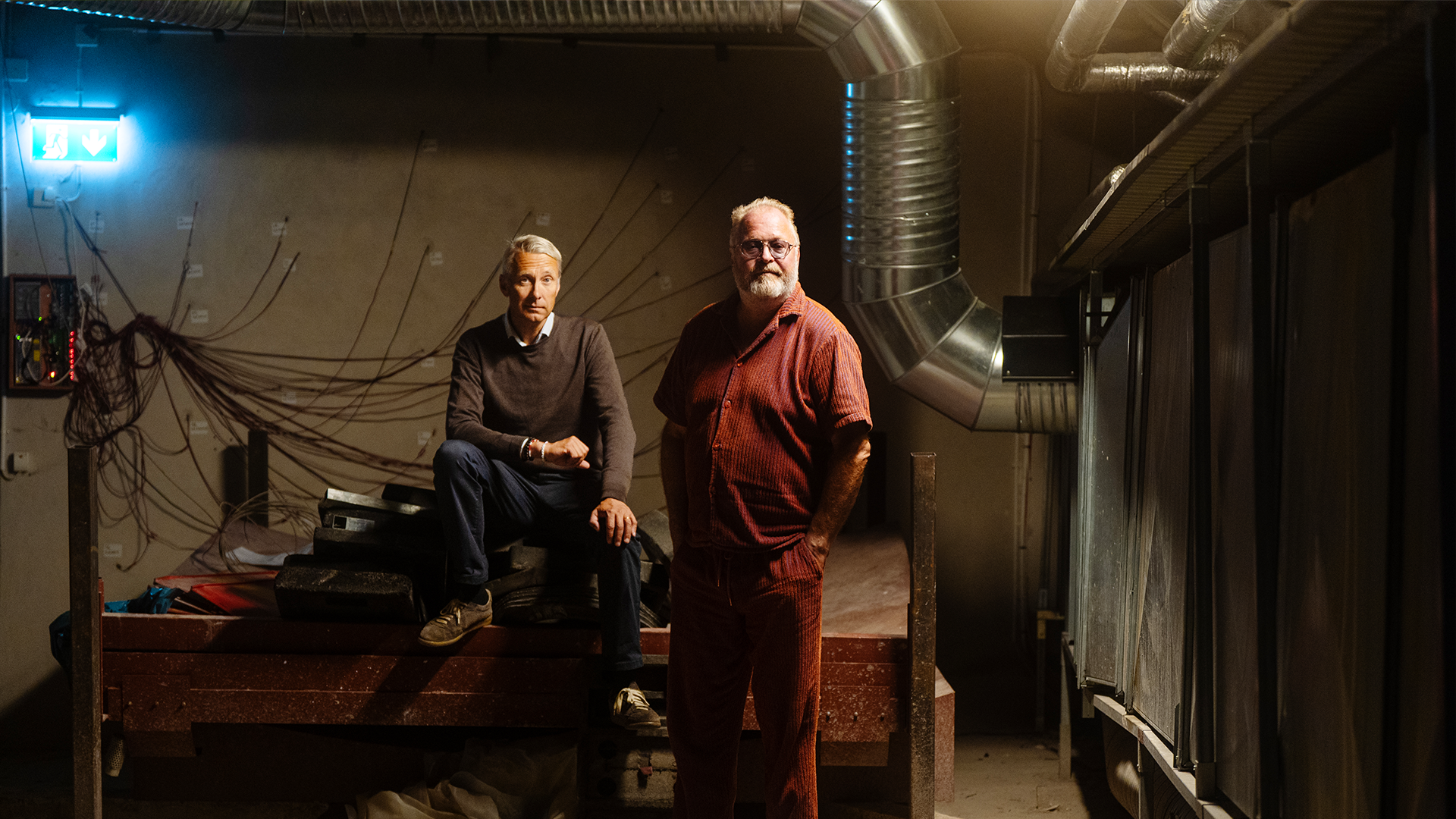 Jan Broman och David von Schinkel blir fotograferade i källaren där Dino ska ta plats. David sitter på en bänk och Jan står bredvid honom. Båda tittar in i kameran.