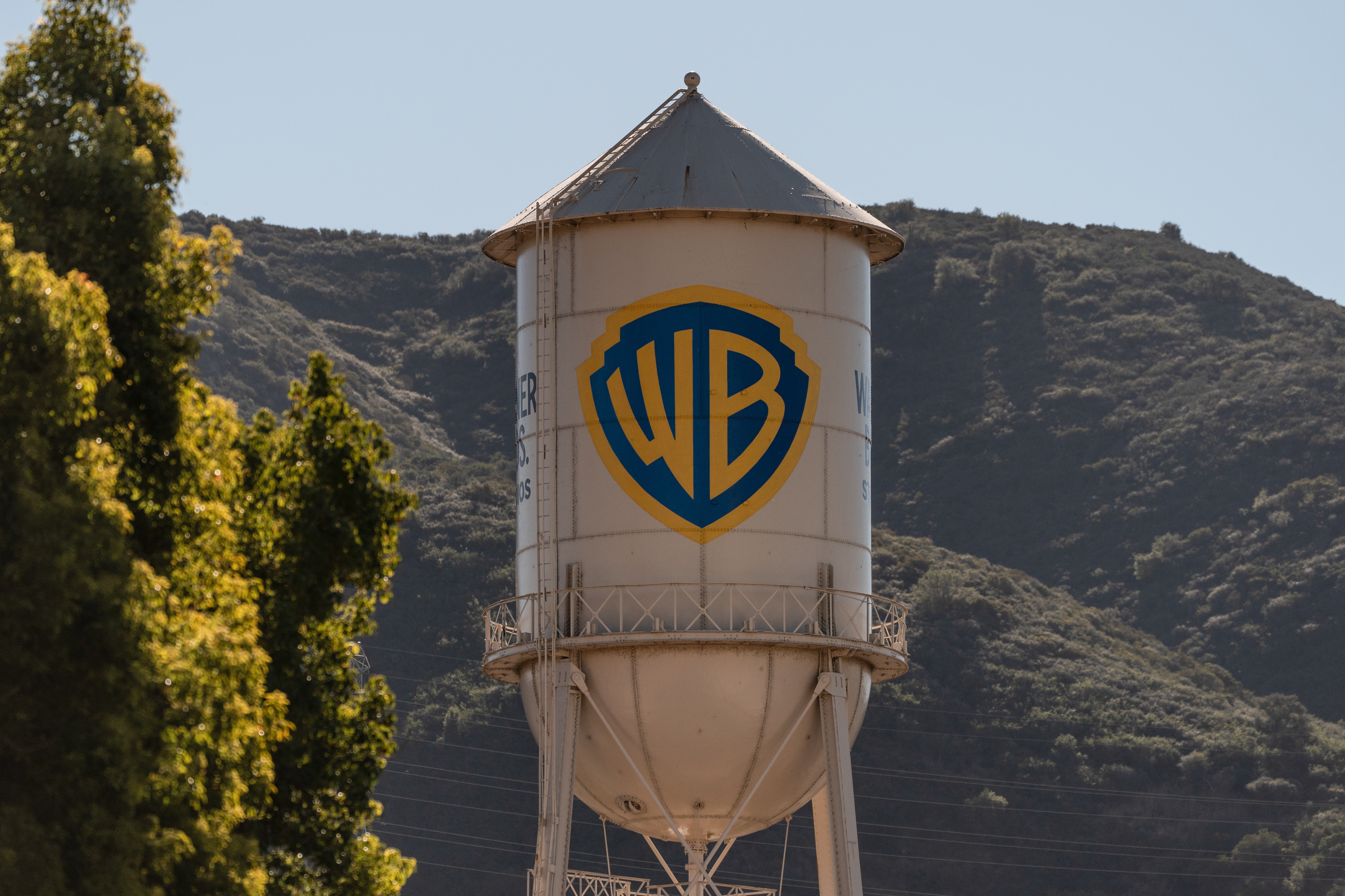 En ikonisk vattentorn med Warner Bros. logotyp - en blå och gul sköld - målat på sidan. Tornet är vitt med en konisk topp och står framför en gräsbevuxen bergssluttning. Gröna träd syns till vänster om tornet under en klar blå himmel.