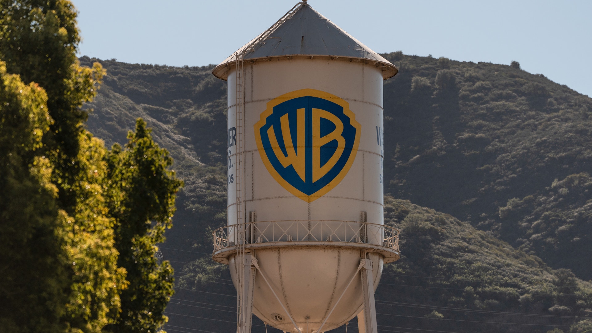 En ikonisk vattentorn med Warner Bros. logotyp - en blå och gul sköld - målat på sidan. Tornet är vitt med en konisk topp och står framför en gräsbevuxen bergssluttning. Gröna träd syns till vänster om tornet under en klar blå himmel.