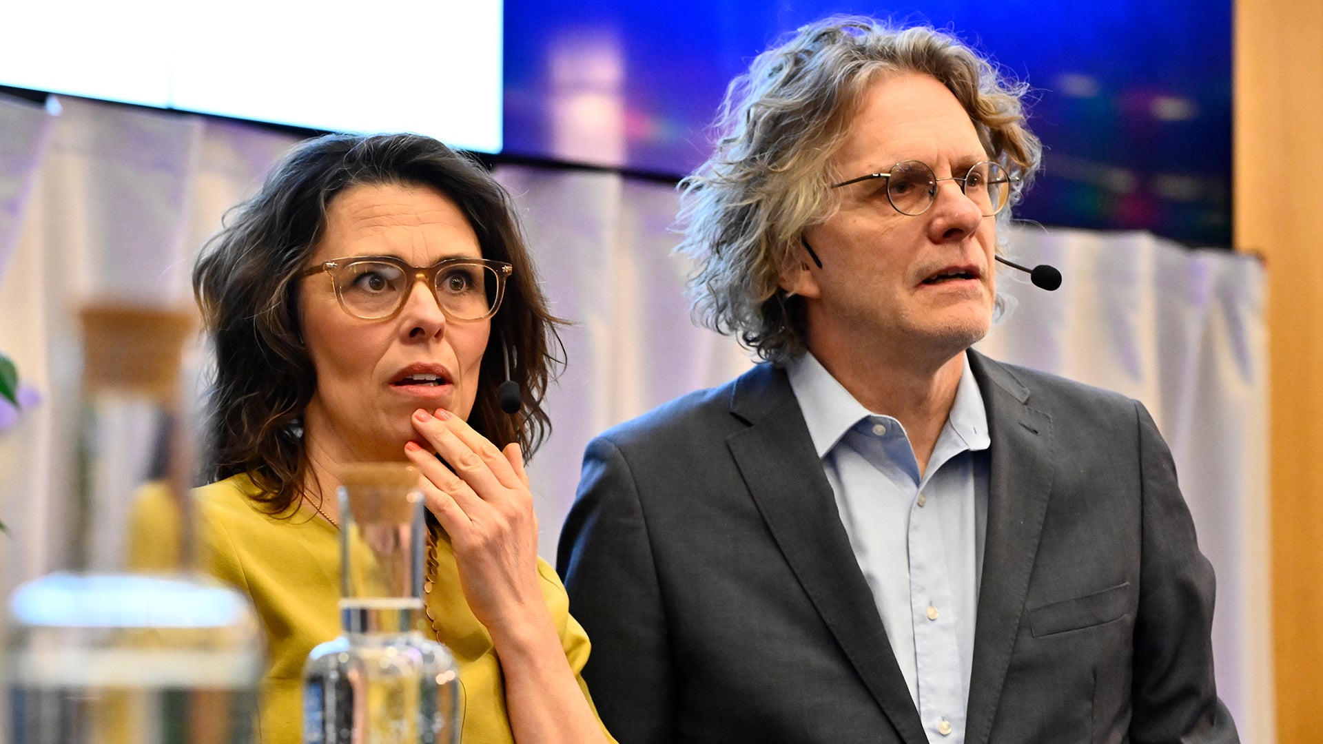 Två personer sitter vid ett bord under en presentation eller konferens. Personen till vänster bär en gul jacka och glasögon, personen till höger bär en grå kavaj. En stor skärm syns i bakgrunden.