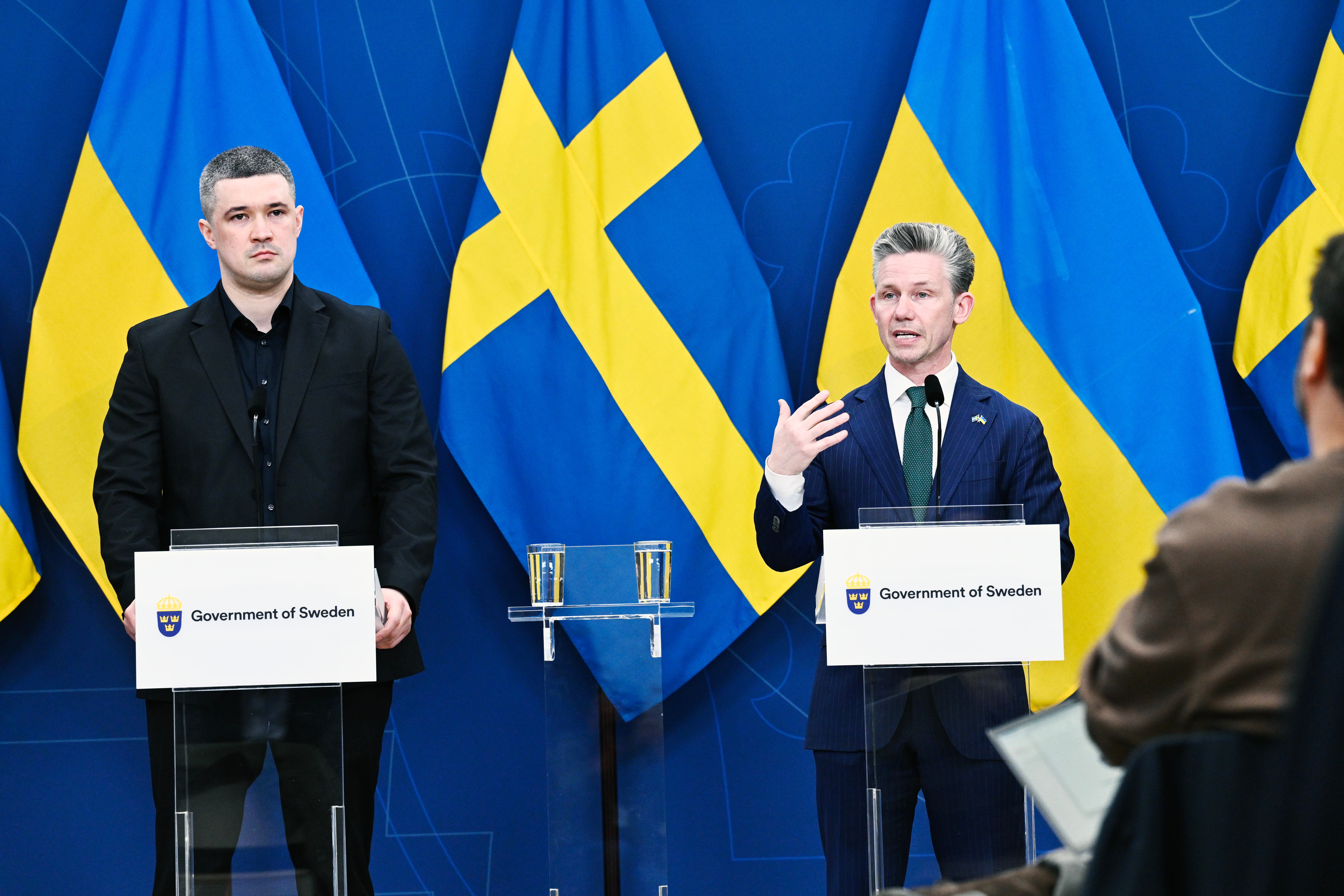 Två män vid podier under en presskonferens framför svenska flaggor med texten 'Government of Sweden'.