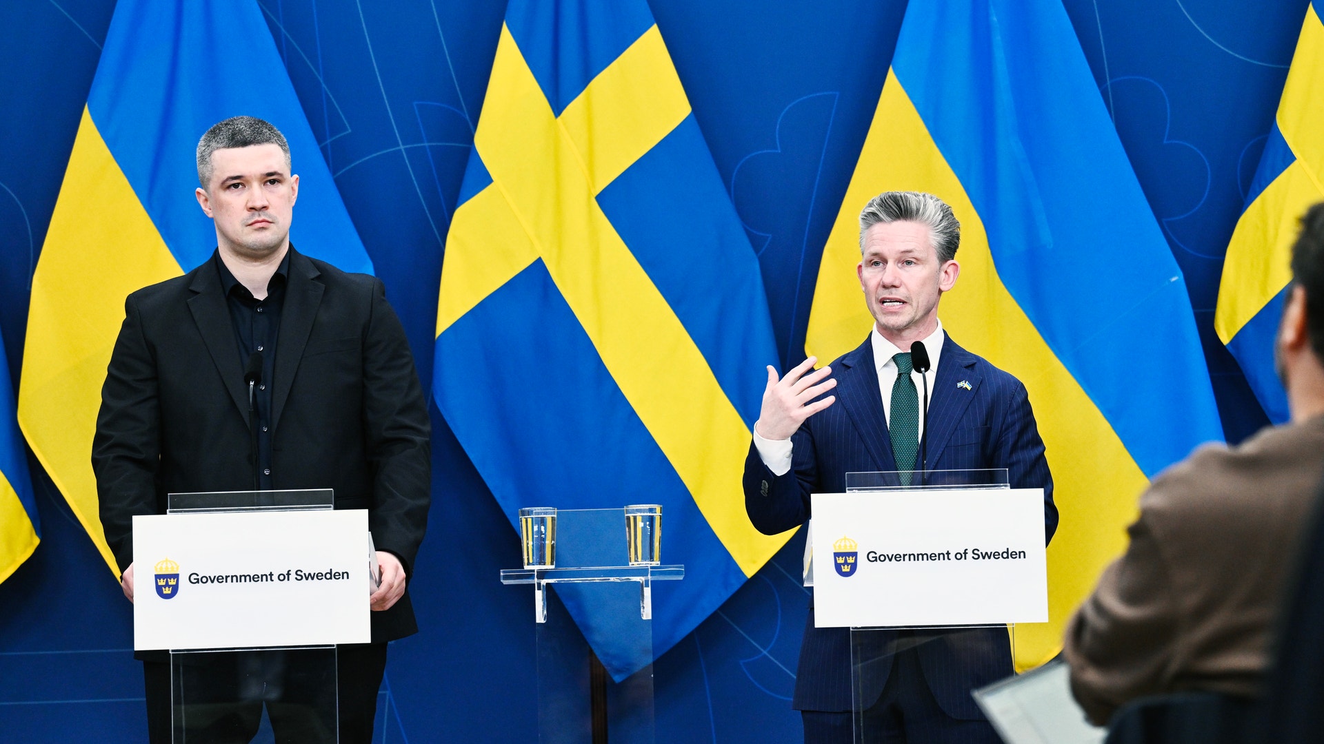 Två män vid podier under en presskonferens framför svenska flaggor med texten 'Government of Sweden'.