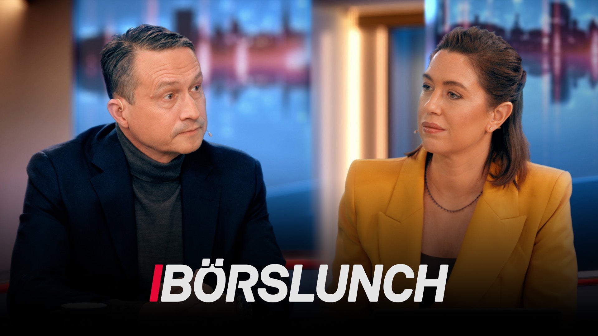 Två personer i diskussion under en tv-sändning med stadsutsikt i bakgrunden.