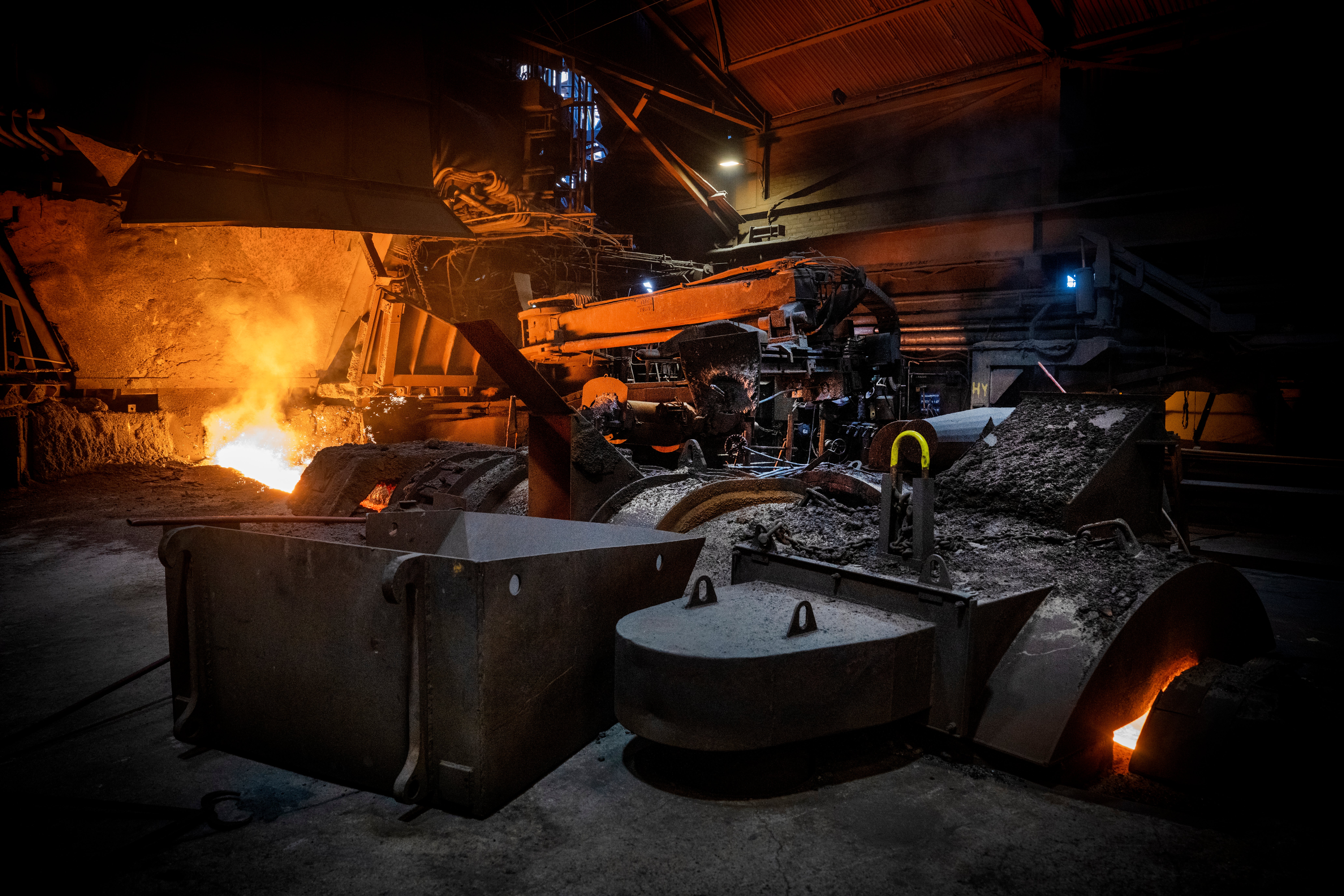 Industriell stålgjuteri med glödande smält metall, stora metallbehållare och grävmaskineri belyst av intensiv värme från produktionsprocessen.