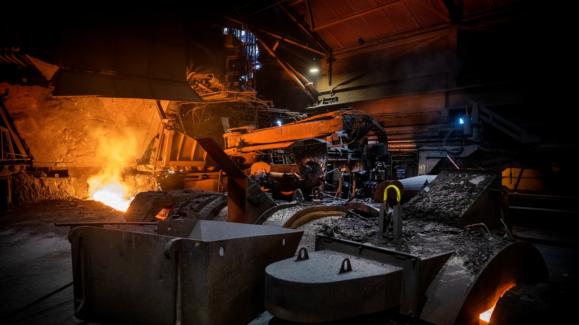 Industriell stålgjuteri med glödande smält metall, stora metallbehållare och grävmaskineri belyst av intensiv värme från produktionsprocessen.