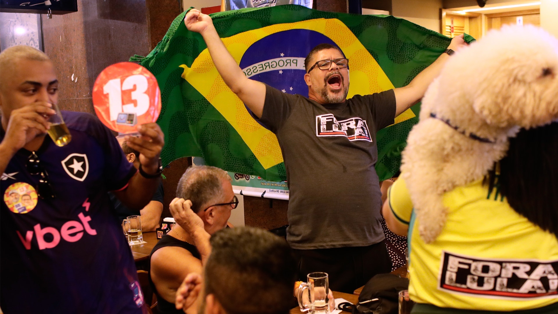 Glada människor firar på en bar, med en brasiliansk flagga och politiska skyltar i bakgrunden.