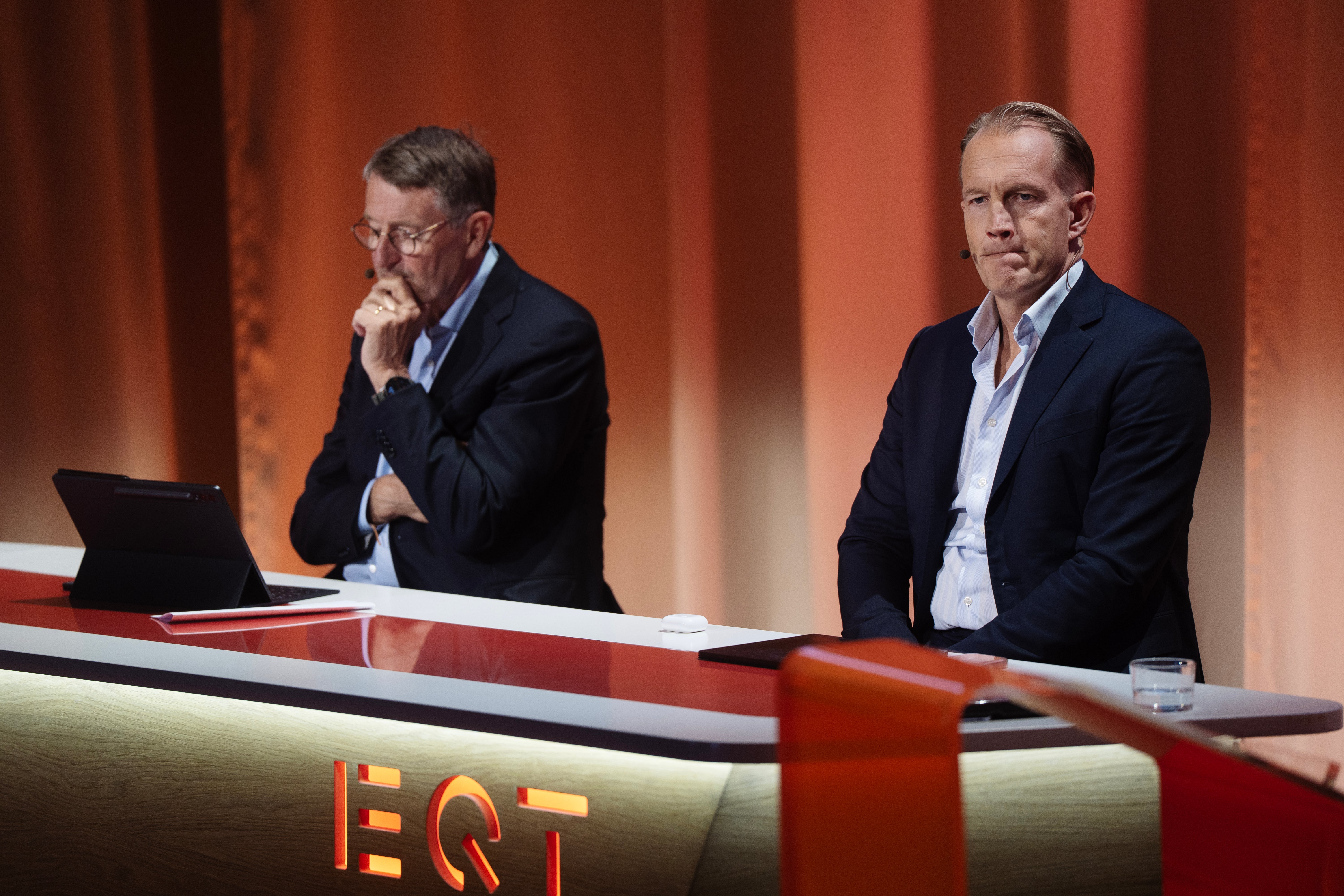 Två män i business-kläder sitter vid ett bord under en paneldiskussion. En man tittar ner medan den andra ser framåt. En laptop och ett glas vatten står på bordet framför dem. Bakgrunden är röd och en EQT-skylt är synlig längst ner.