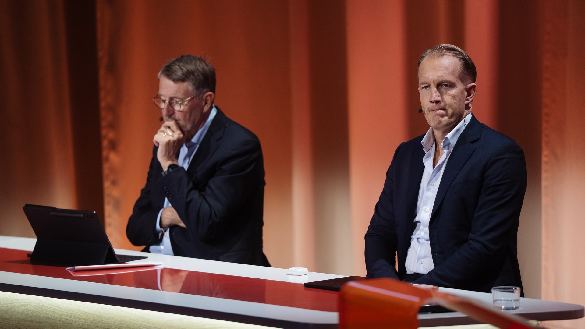 Två män i business-kläder sitter vid ett bord under en paneldiskussion. En man tittar ner medan den andra ser framåt. En laptop och ett glas vatten står på bordet framför dem. Bakgrunden är röd och en EQT-skylt är synlig längst ner.