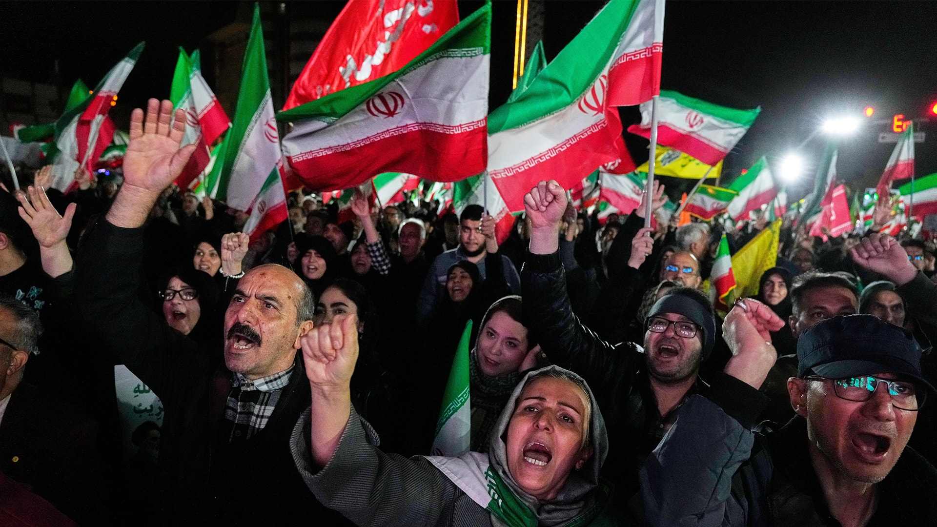 En folkmängd på natten som viftar med iranska flaggor och höjer händerna.