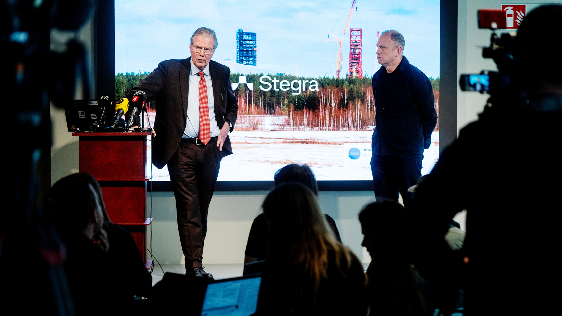 Två män presenterar på en scen under en konferens med publik och en stor skärm med Stegra-logotypen i bakgrunden.