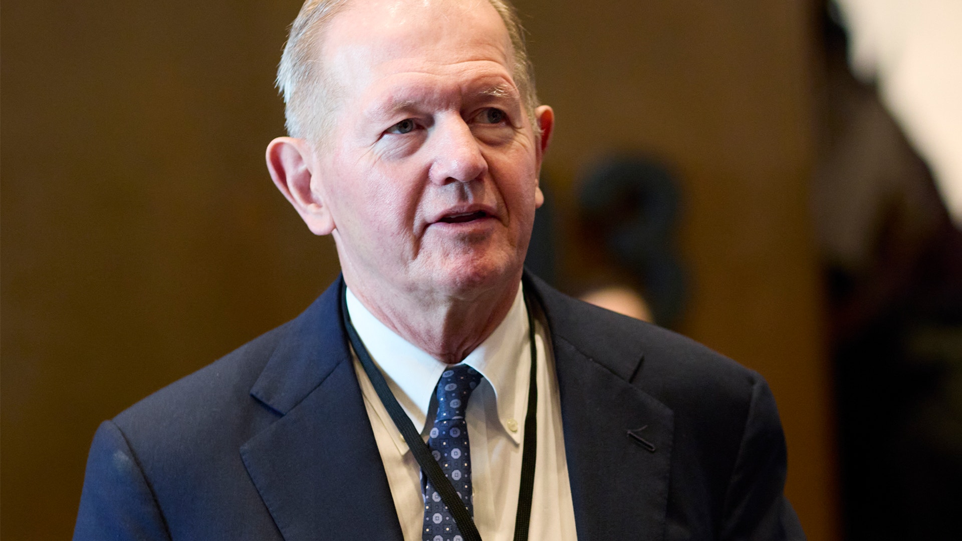 En äldre man i mörk kostym och slips framför en neutral bakgrund.