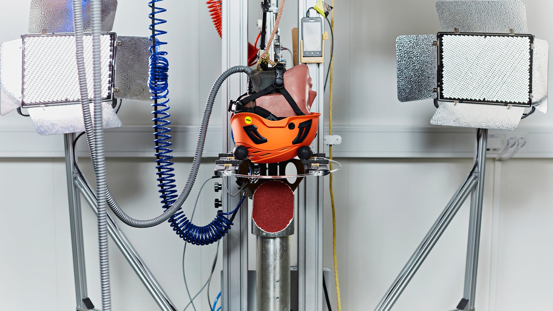 En 3D-skrivare med orange och svart skrivarhuvud monterad på en metallram, omgiven av organiserade slangar och kablar på väggen.