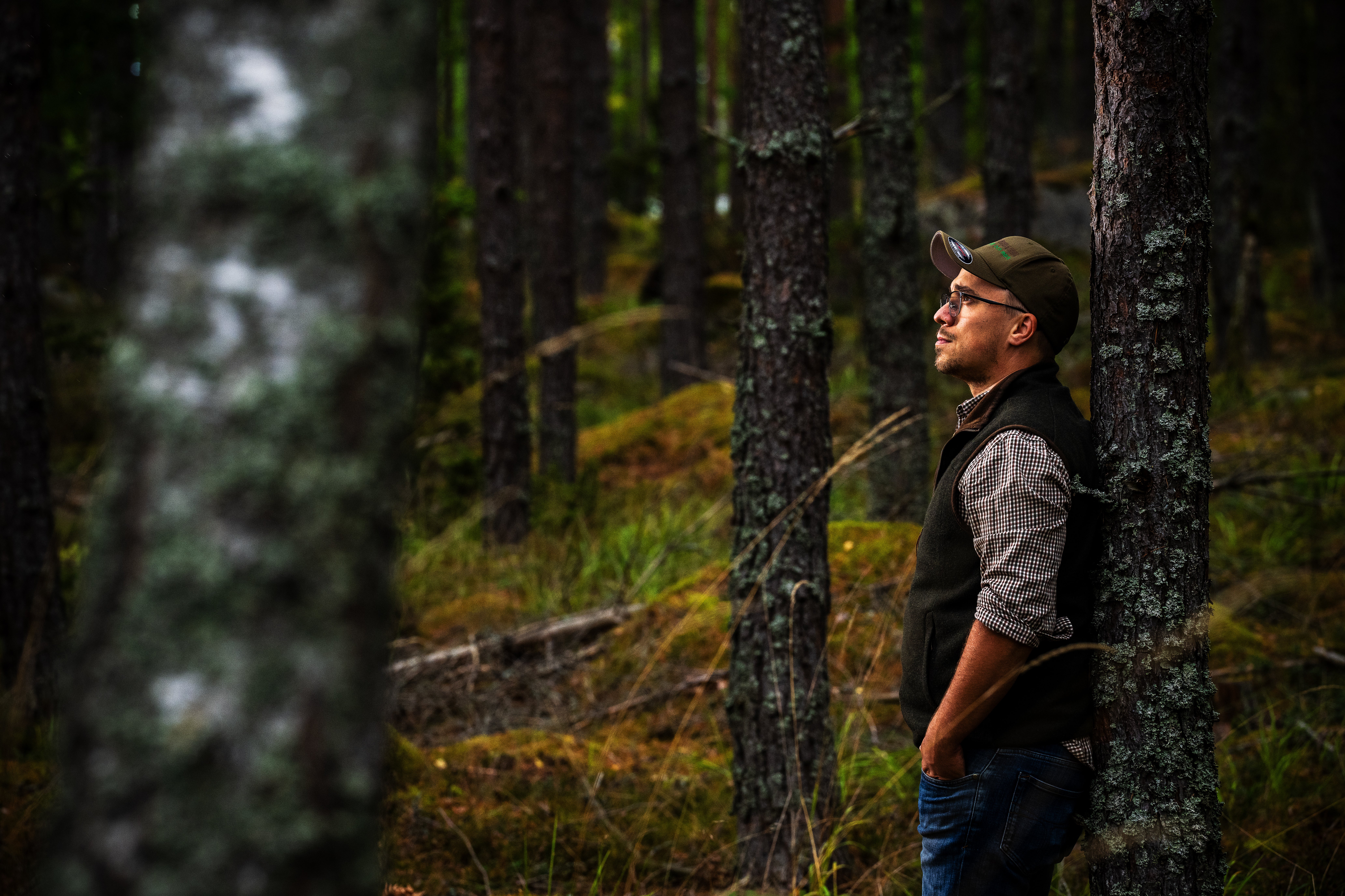 En man lutar sig mot ett träd i en skog och blickar ut mellan trädstammarna.