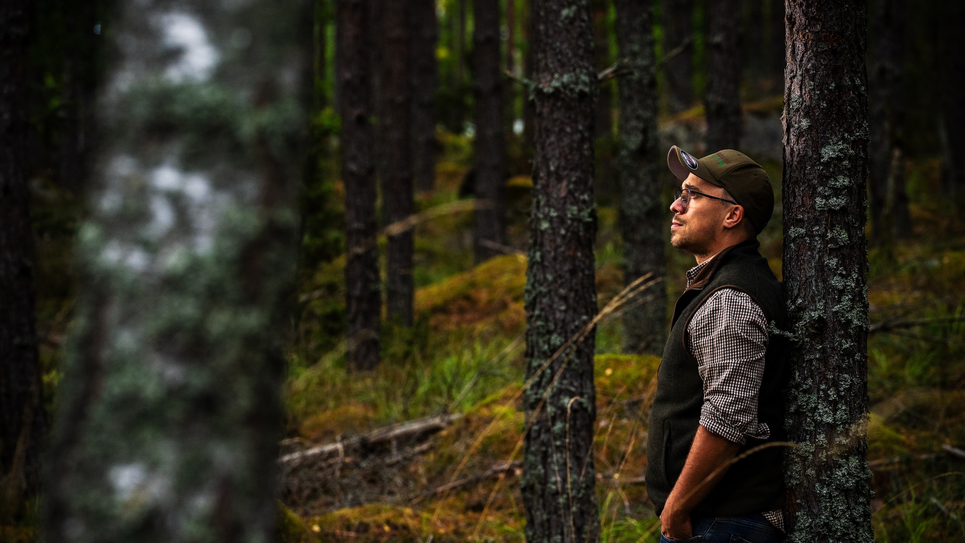 En man lutar sig mot ett träd i en skog och blickar ut mellan trädstammarna.