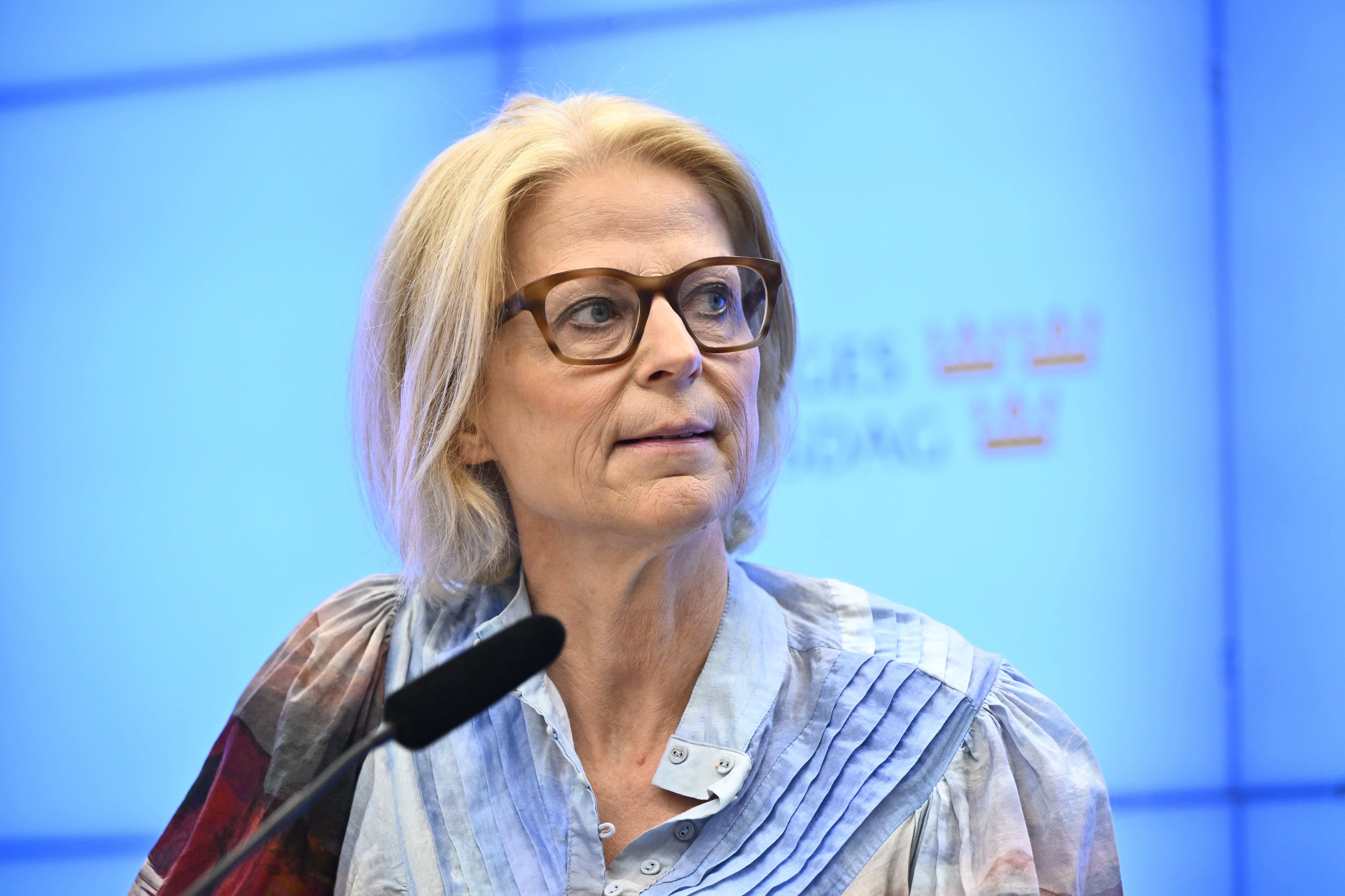Porträtt av en äldre kvinna med blont hår och glasögon mot en blå bakgrund.