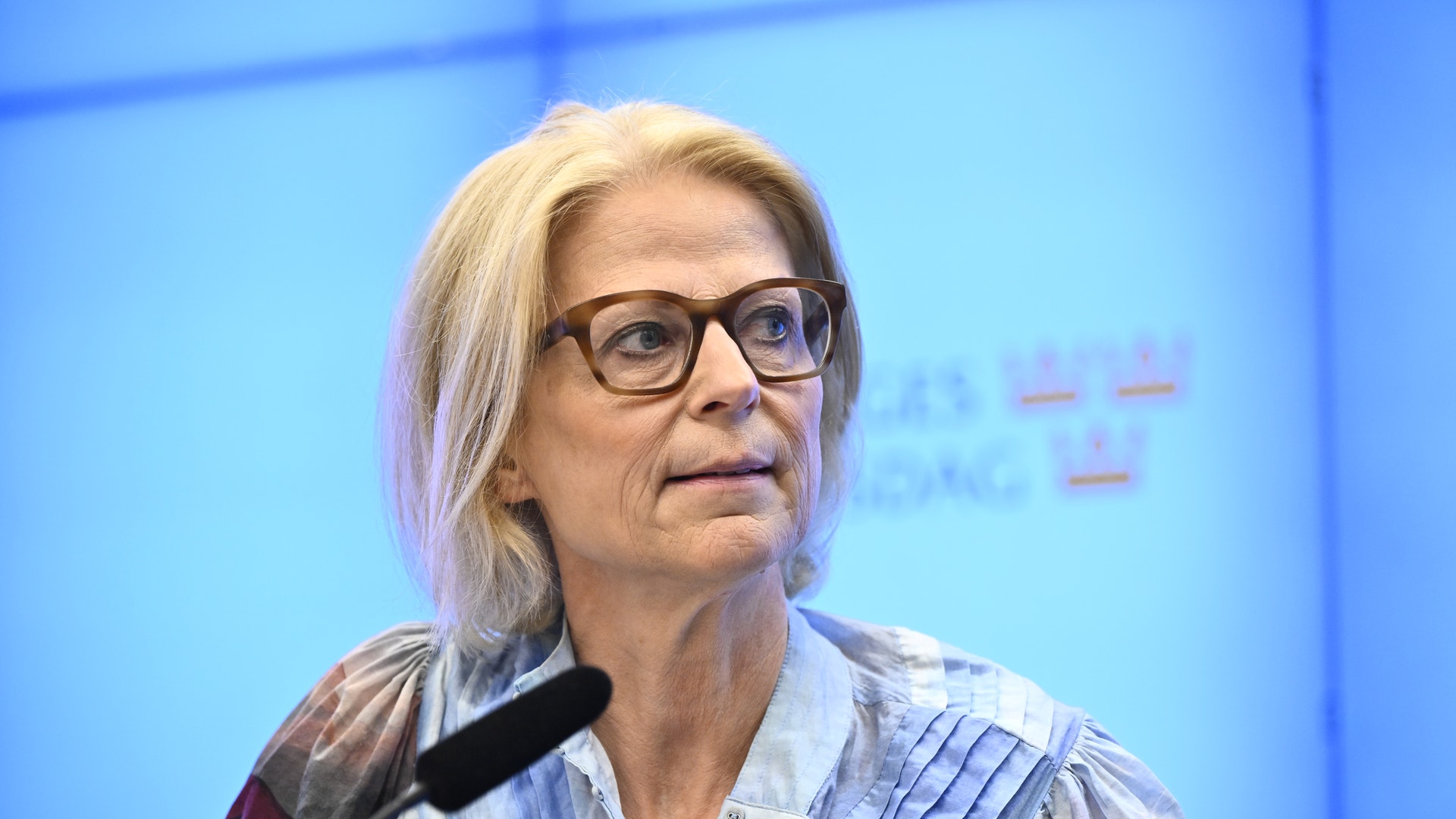 Porträtt av en äldre kvinna med blont hår och glasögon mot en blå bakgrund.