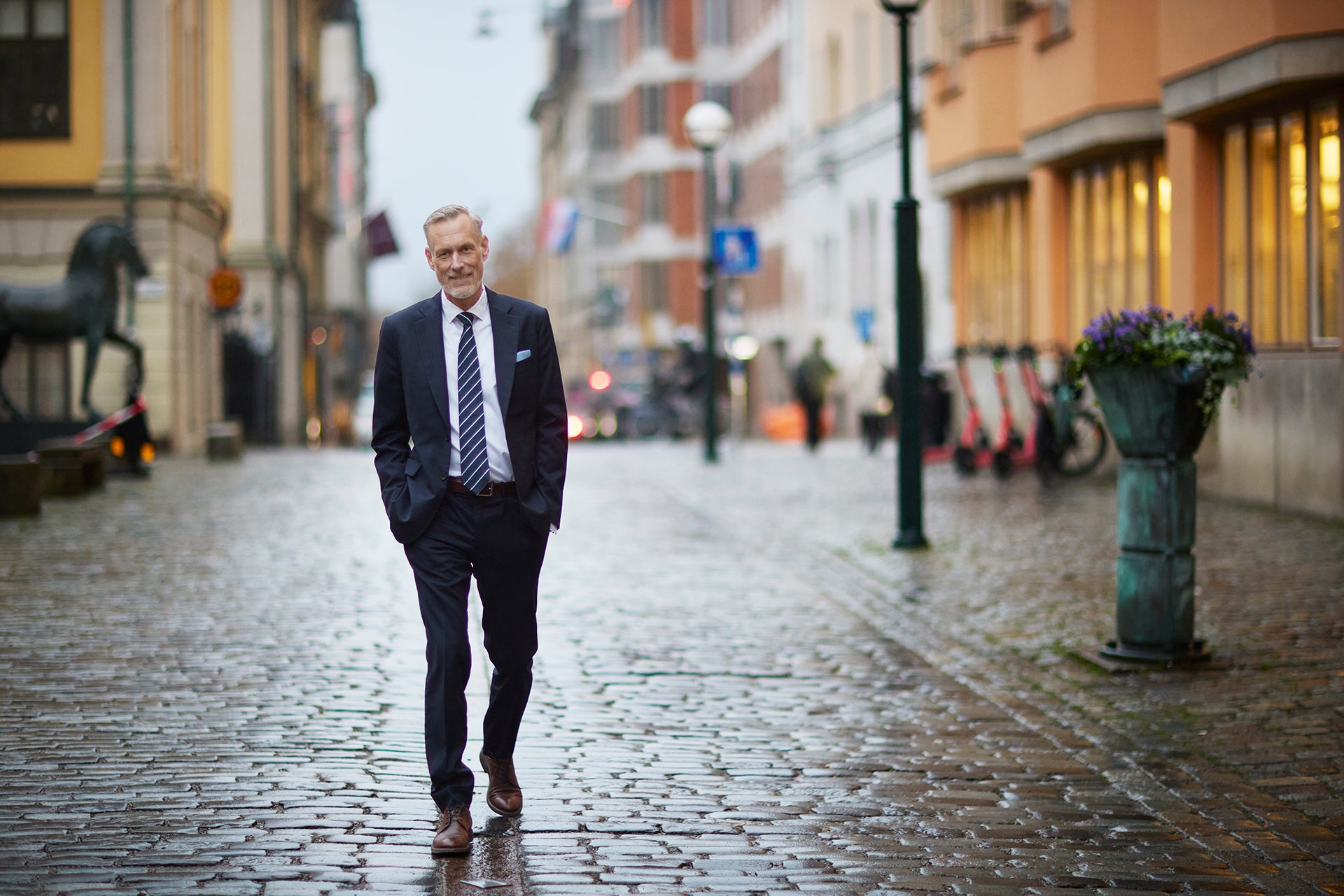 En man i mörk kostym och slips går längs en kullerstensgata i en stad.