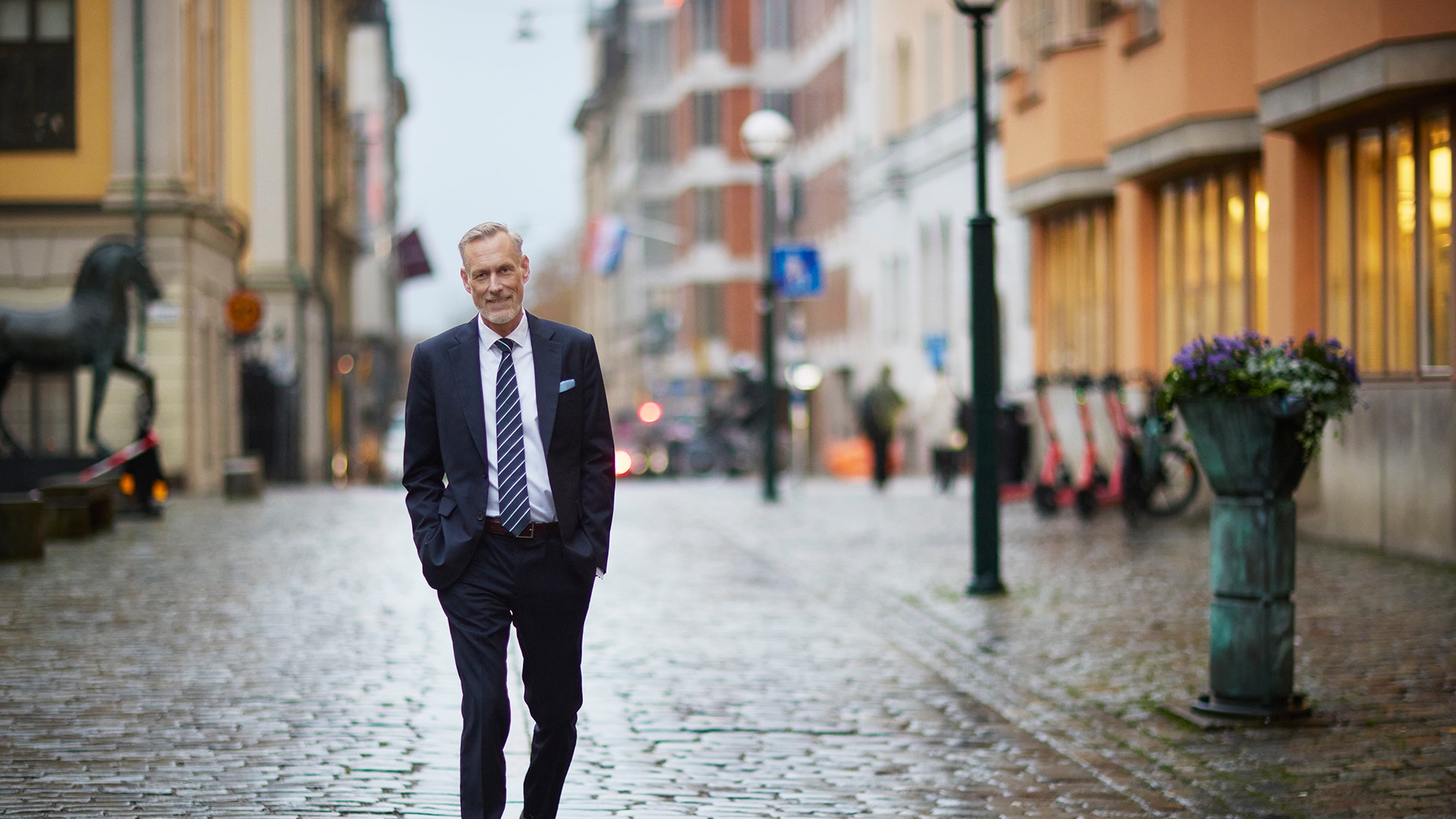 En man i mörk kostym och slips går längs en kullerstensgata i en stad.