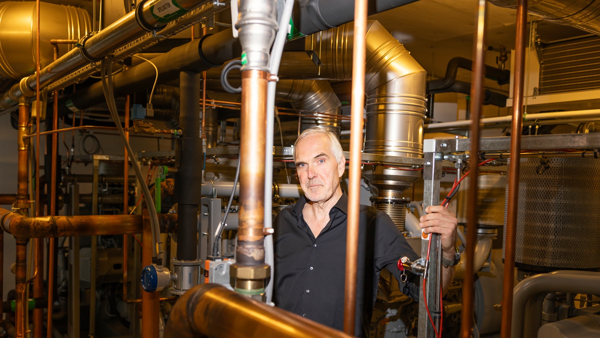 Man i mörk skjorta står bland industriell utrustning med kopparrör och metallrör.