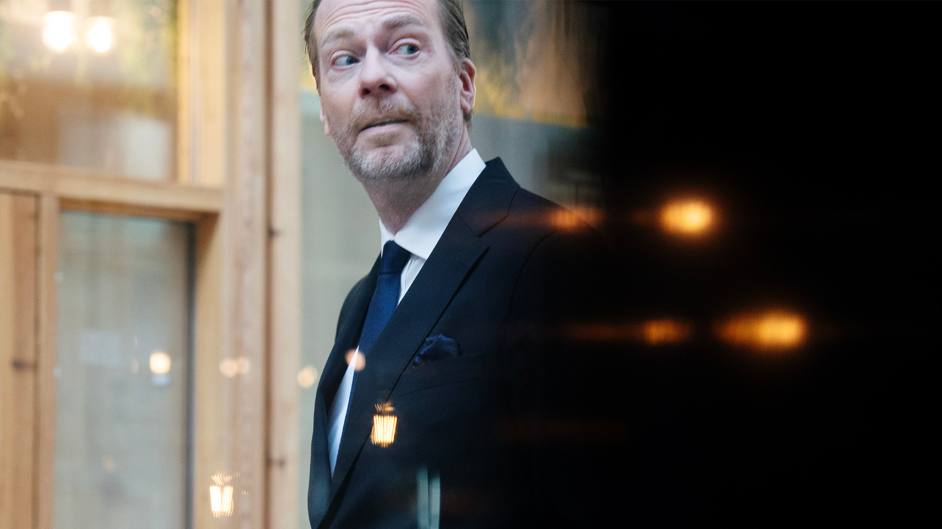 Porträtt av en man i mörk kostym och slips framför en elegant interiör med belysning.