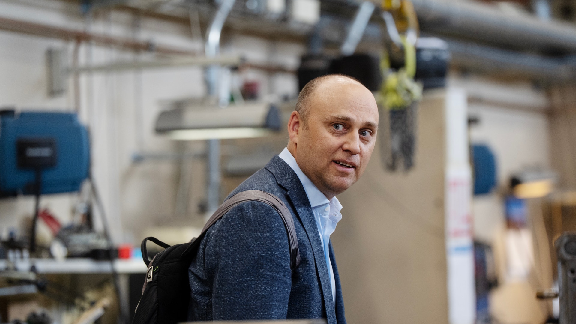 En man i mörkblå kavaj och ljusblå skjorta står i en fabrik och ser över axeln mot kameran.