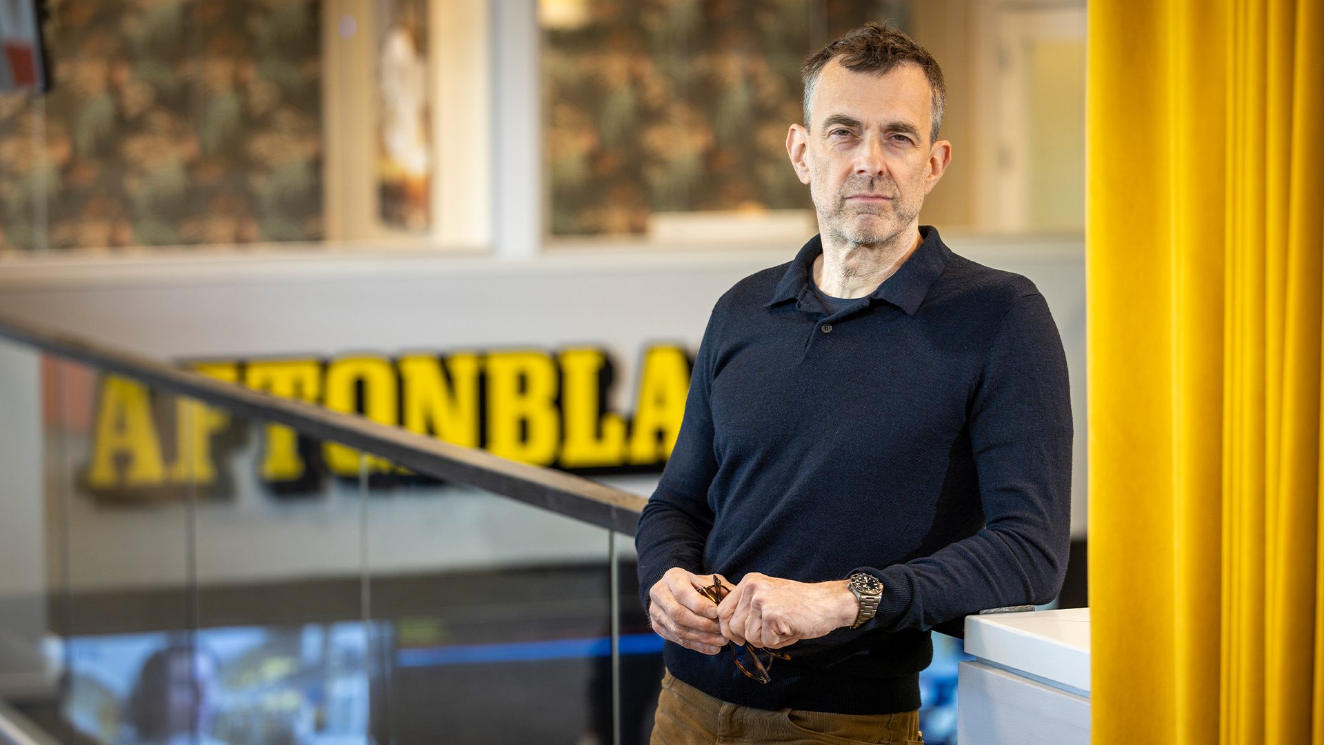 Porträtt av en man i mörk tröja som står framför en vägg med gul dekoration i en professionell miljö.
