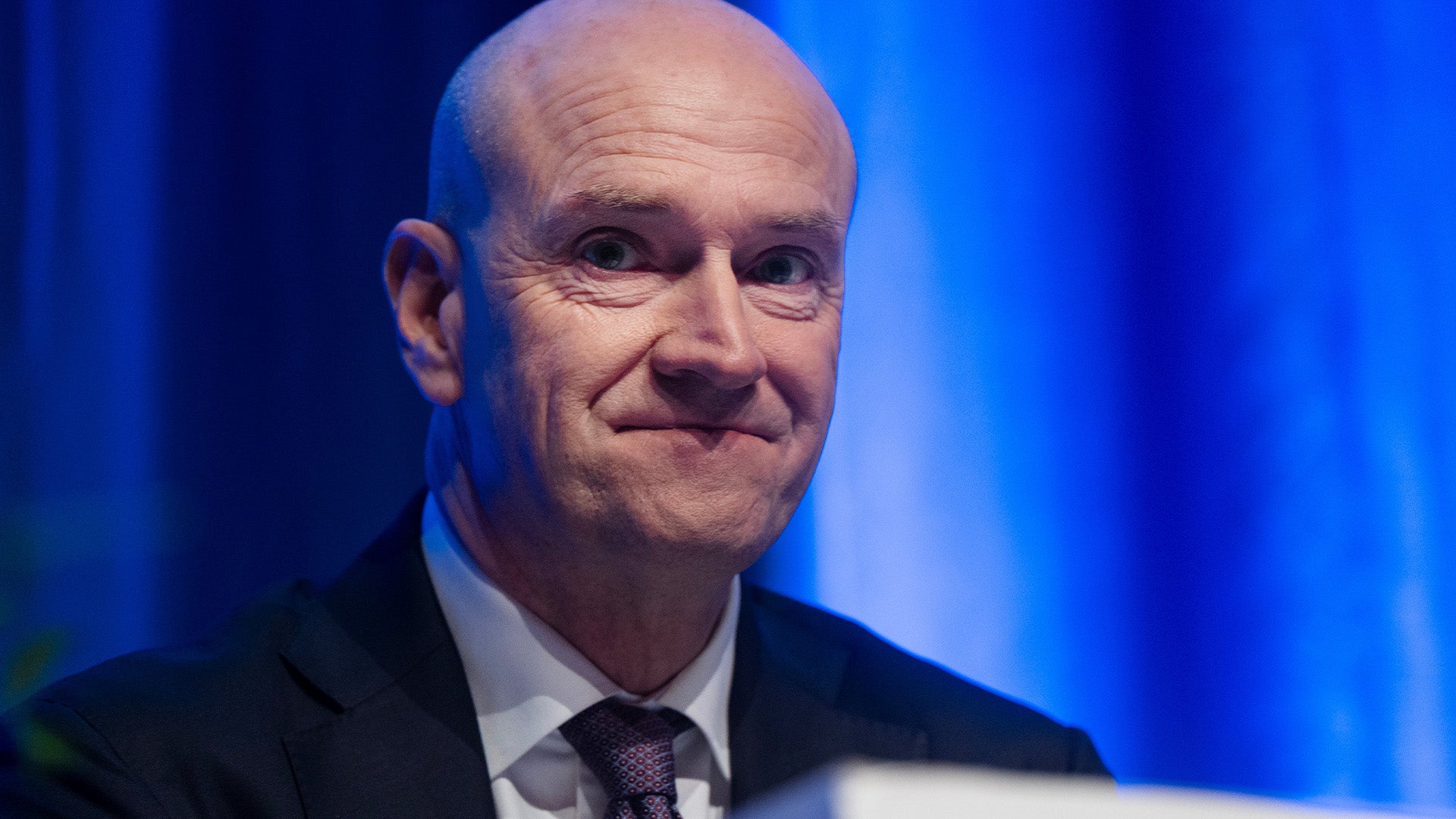 En man i kostym och slips framför en blå bakgrund.