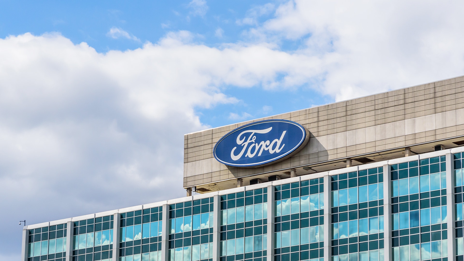 Ford-logotyp på taket av en modern kontorsbyggnad med glasfönster och blå himmel med vita moln.