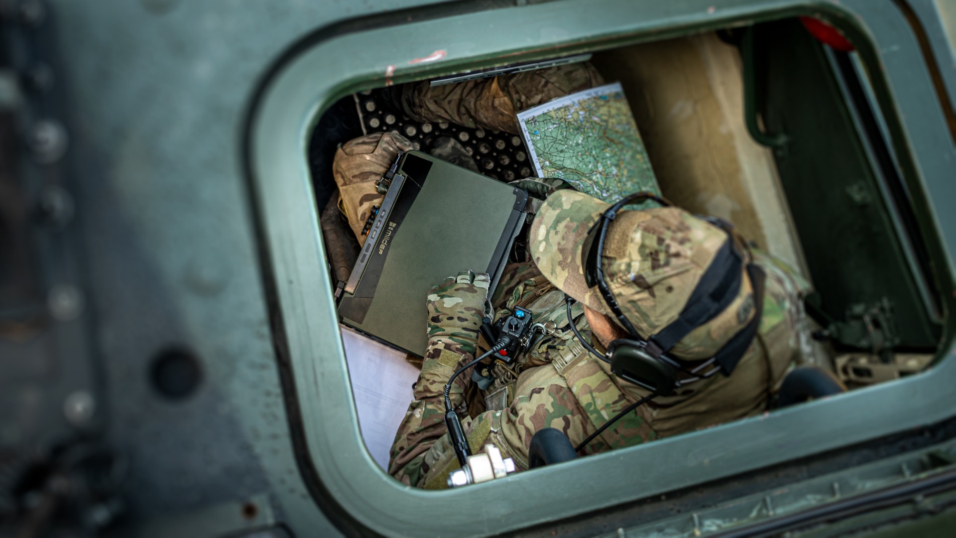 Soldat i militär uniform och utrustning sedd uppifrån genom en öppen lucka på ett militärfordon.