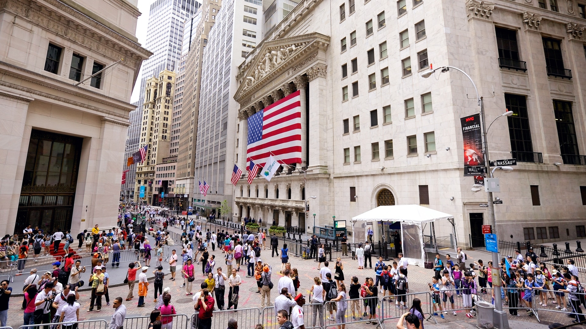 New York Stock Exchange på Wall Street med en stor amerikansk flagga på fasaden. Många människor går på gatan framför byggnaden.