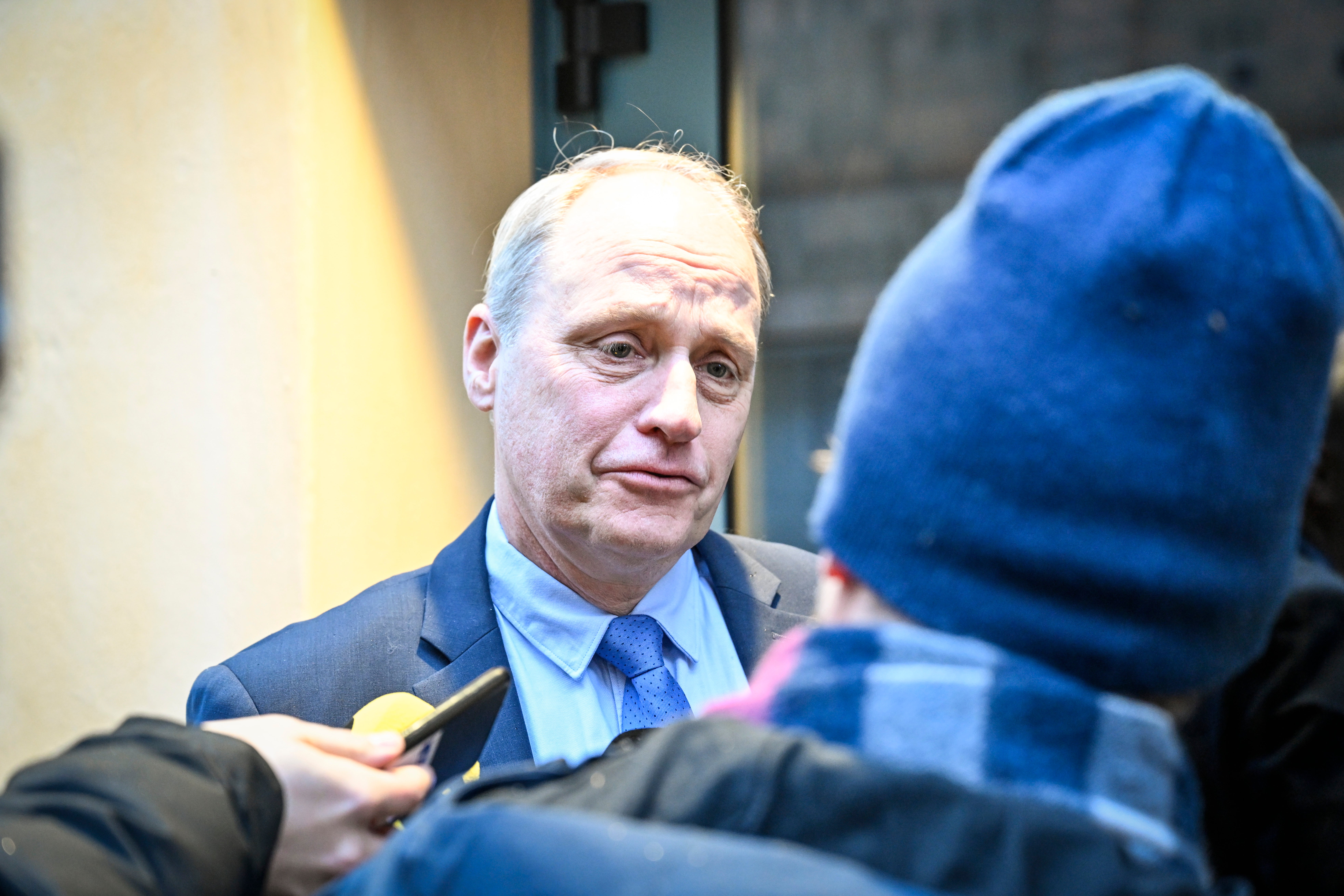 En man i kostym och blå slips intervjuas av en person i blå jacka.