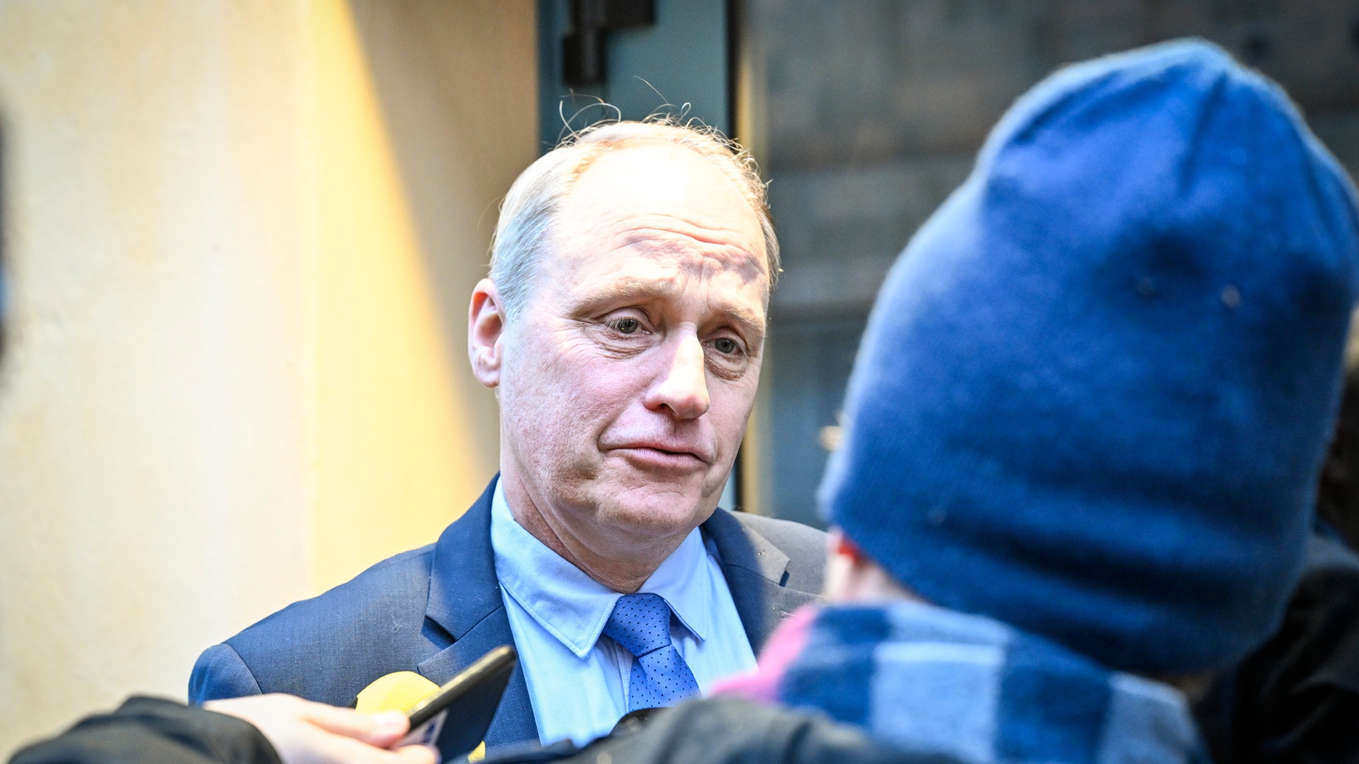 En man i kostym och blå slips intervjuas av en person i blå jacka.