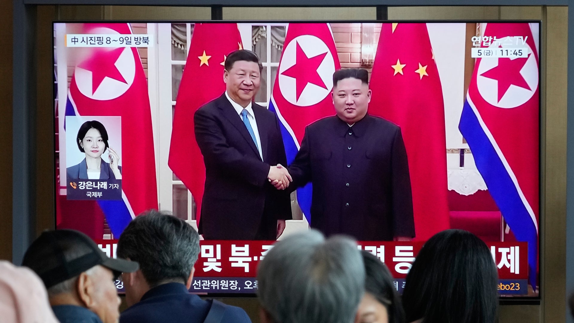 TV-skärm visar två män i mörka kostymer som skakar hand framför röda flaggor med gula stjärnor. Nyhetssändning med koreansk text. Människor i förgrunden tittar på skärmen.