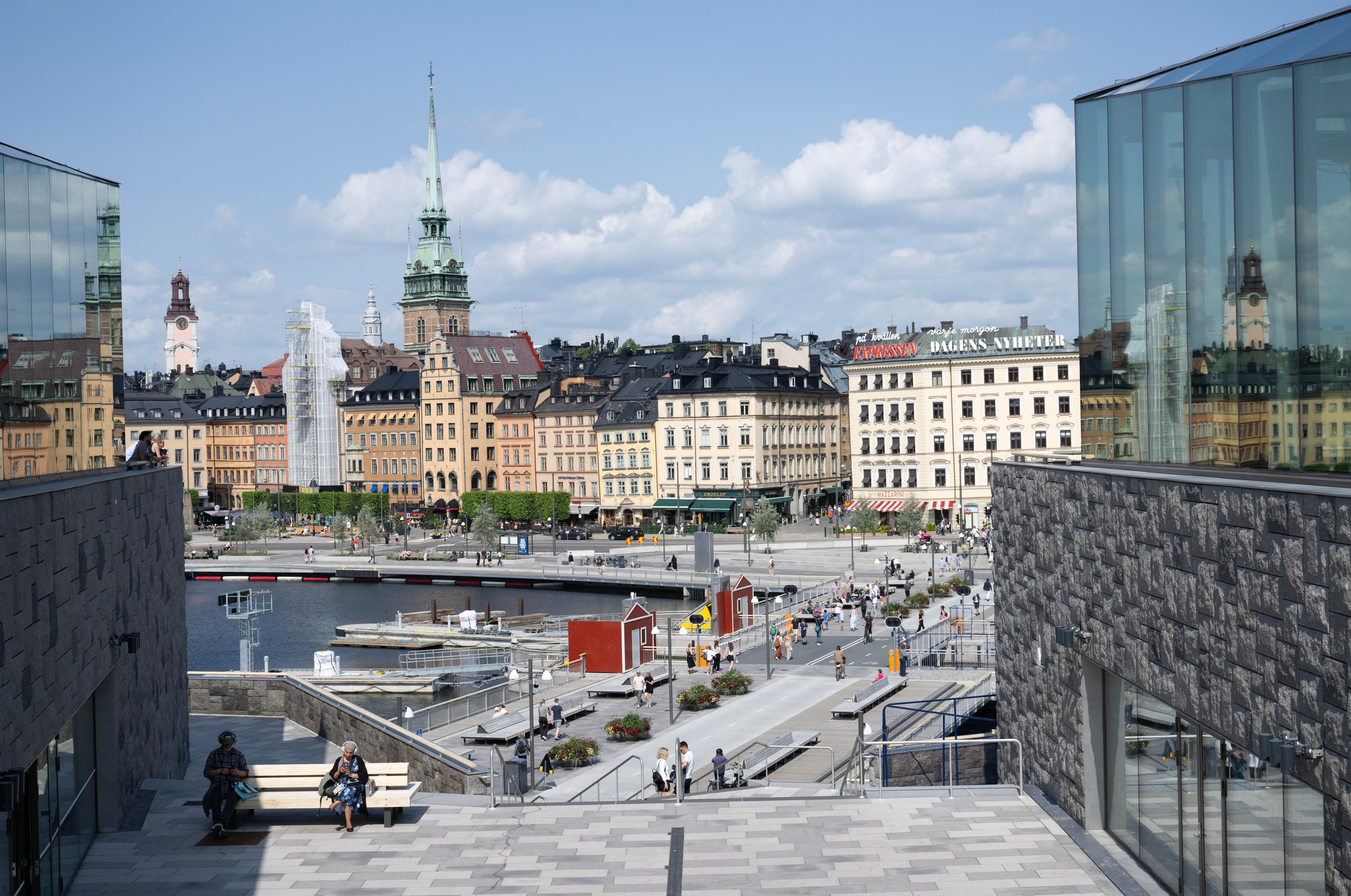 Utsikt över en europeisk stad med färgglada gamla byggnader, en kyrka med grön spira, en kanal och moderna byggnader i förgrunden.