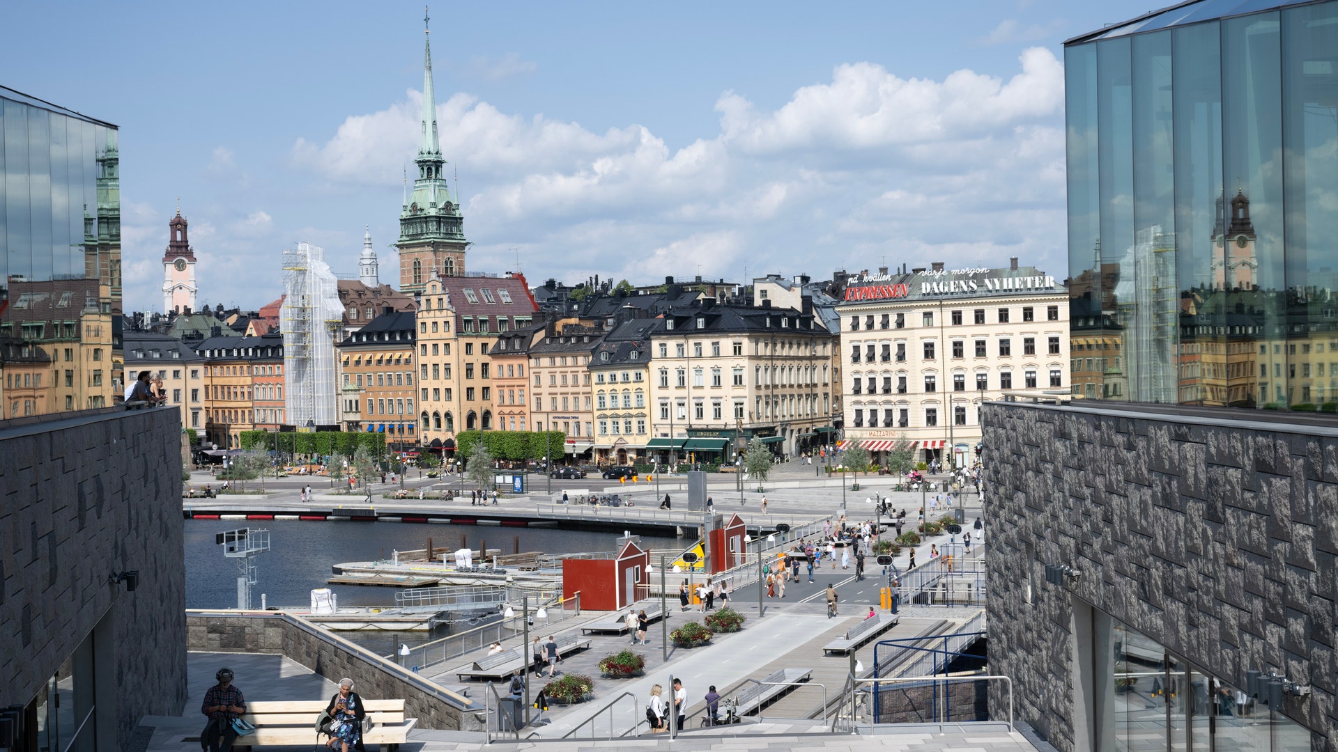 Utsikt över en europeisk stad med färgglada gamla byggnader, en kyrka med grön spira, en kanal och moderna byggnader i förgrunden.