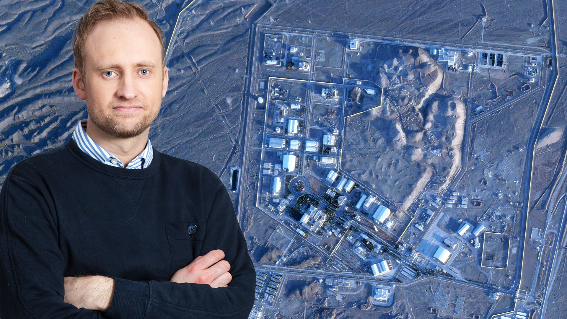 Man i mörkblå tröja framför flygfoto av industrianläggning sedd från ovan.