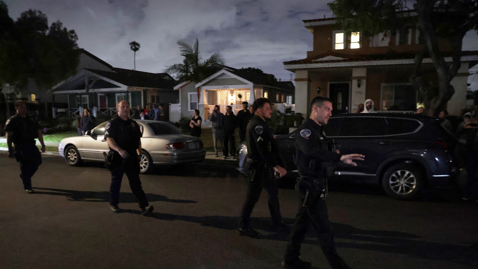 Polisöversikt på en bostadsgata under kvällen med poliser, bilar och belyst bostadshus i bakgrunden.