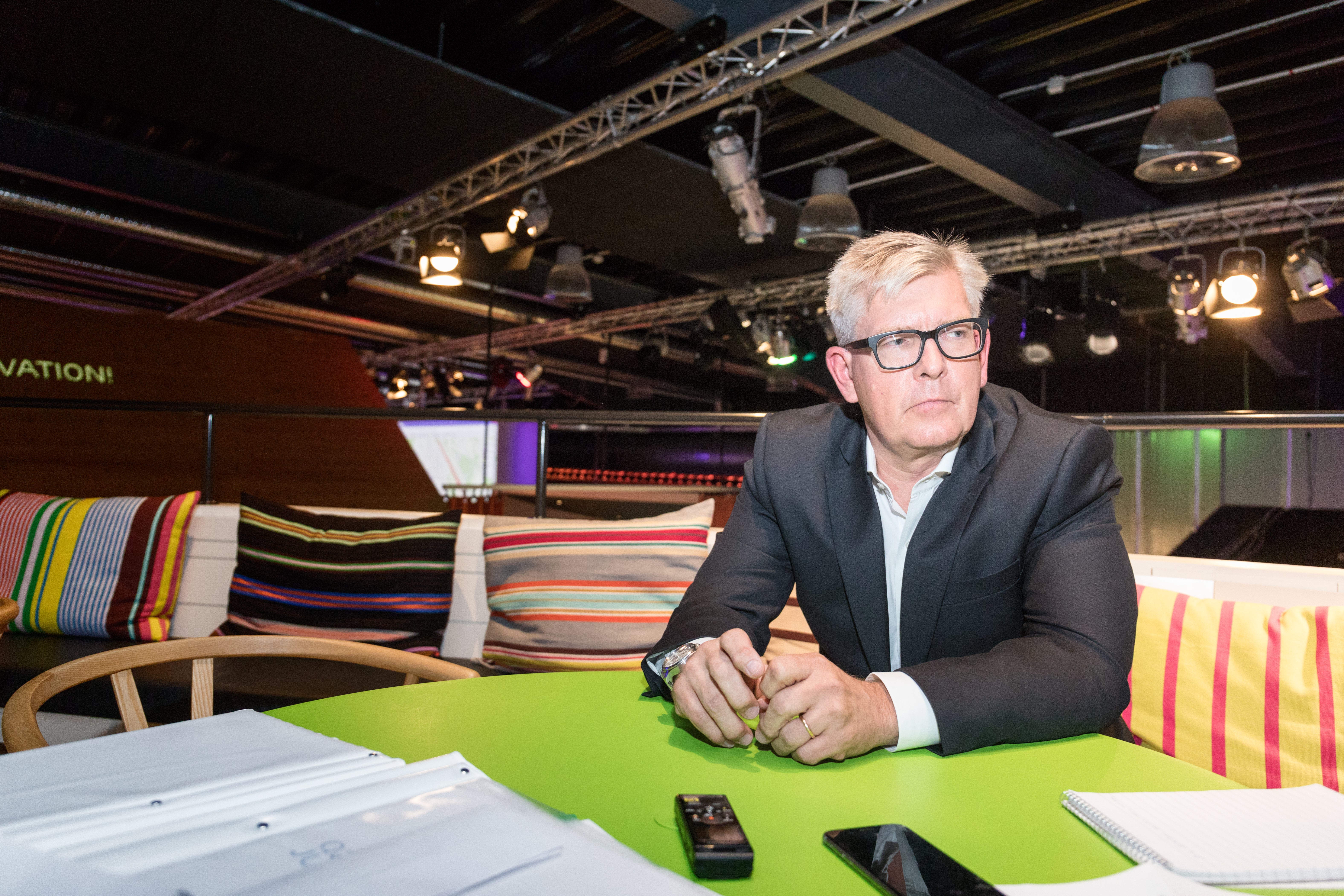 En man med glasögon och grått hår sitter vid ett grönt bord i en modern studiomiljö.