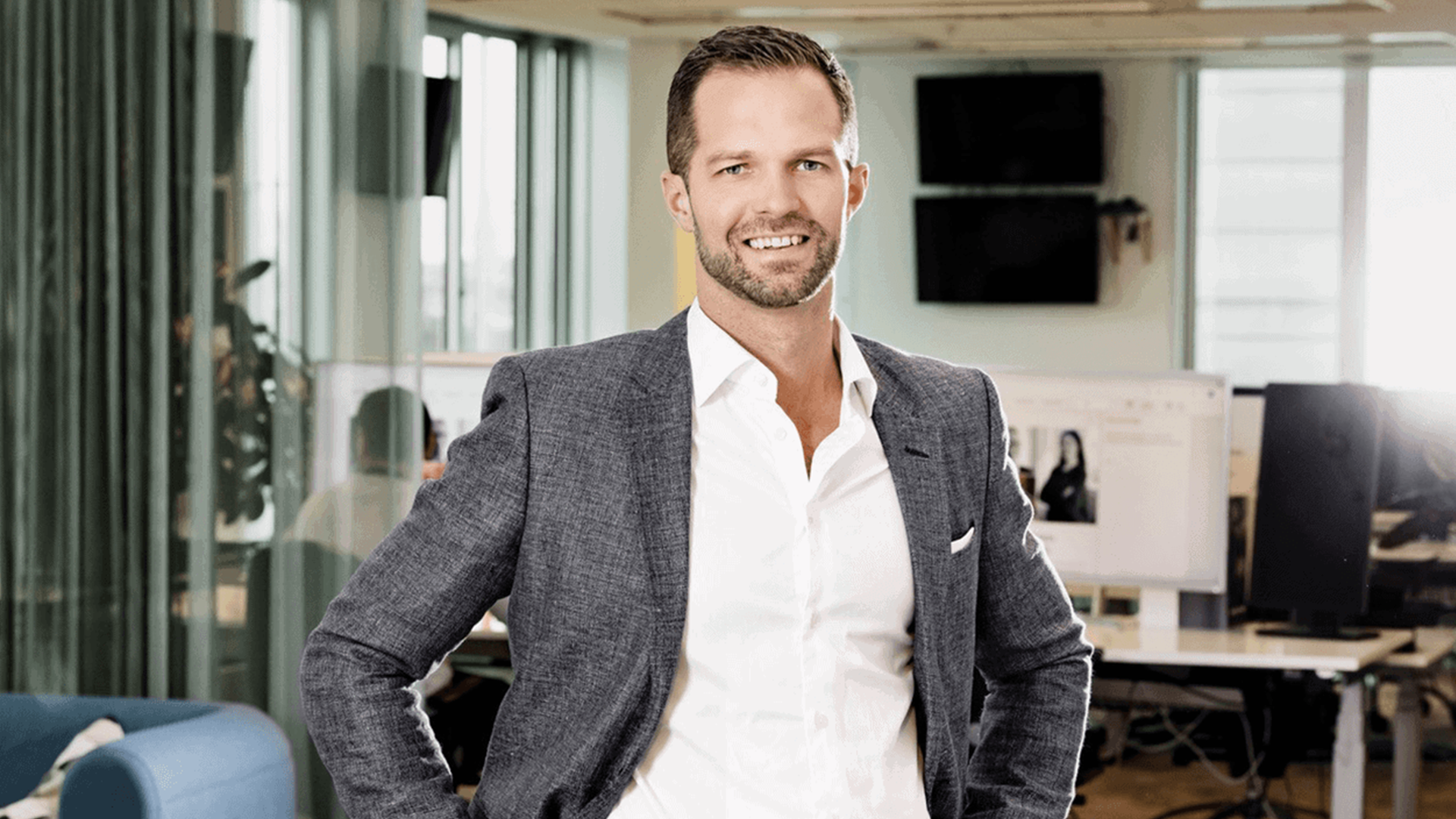 En man i grå blazer och vit skjorta står leende i ett modernt kontor.