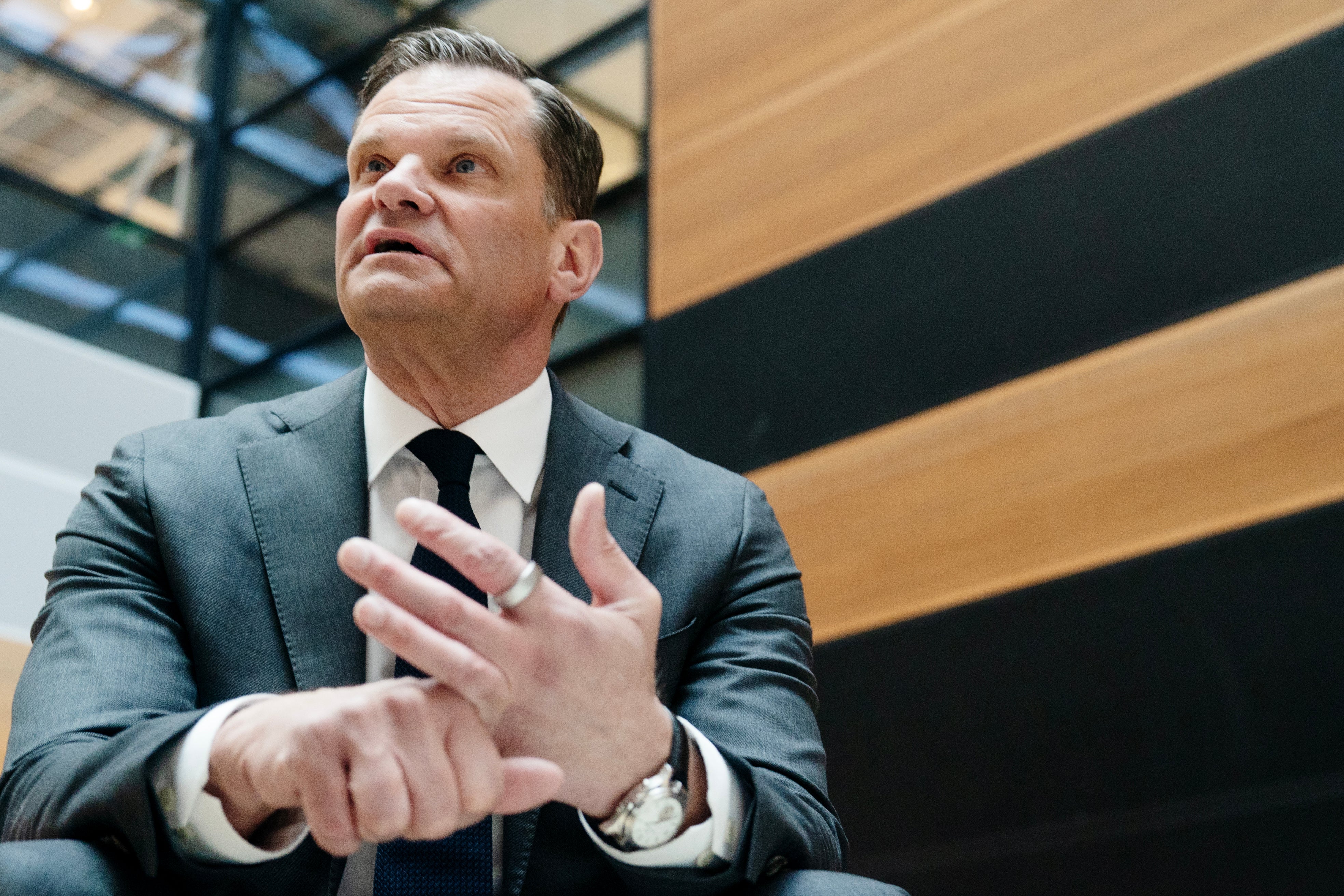 En man i kostym och slips håller ett tal eller presentation med gestikulerande händer framför en modern byggnad.