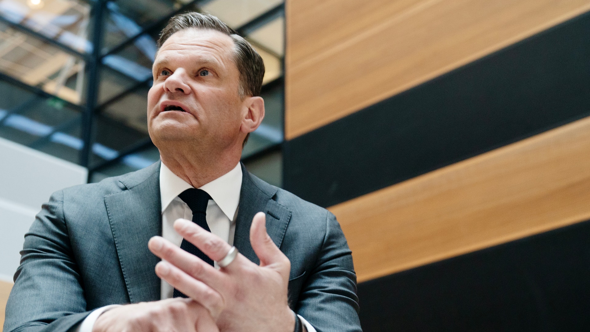 En man i kostym och slips håller ett tal eller presentation med gestikulerande händer framför en modern byggnad.