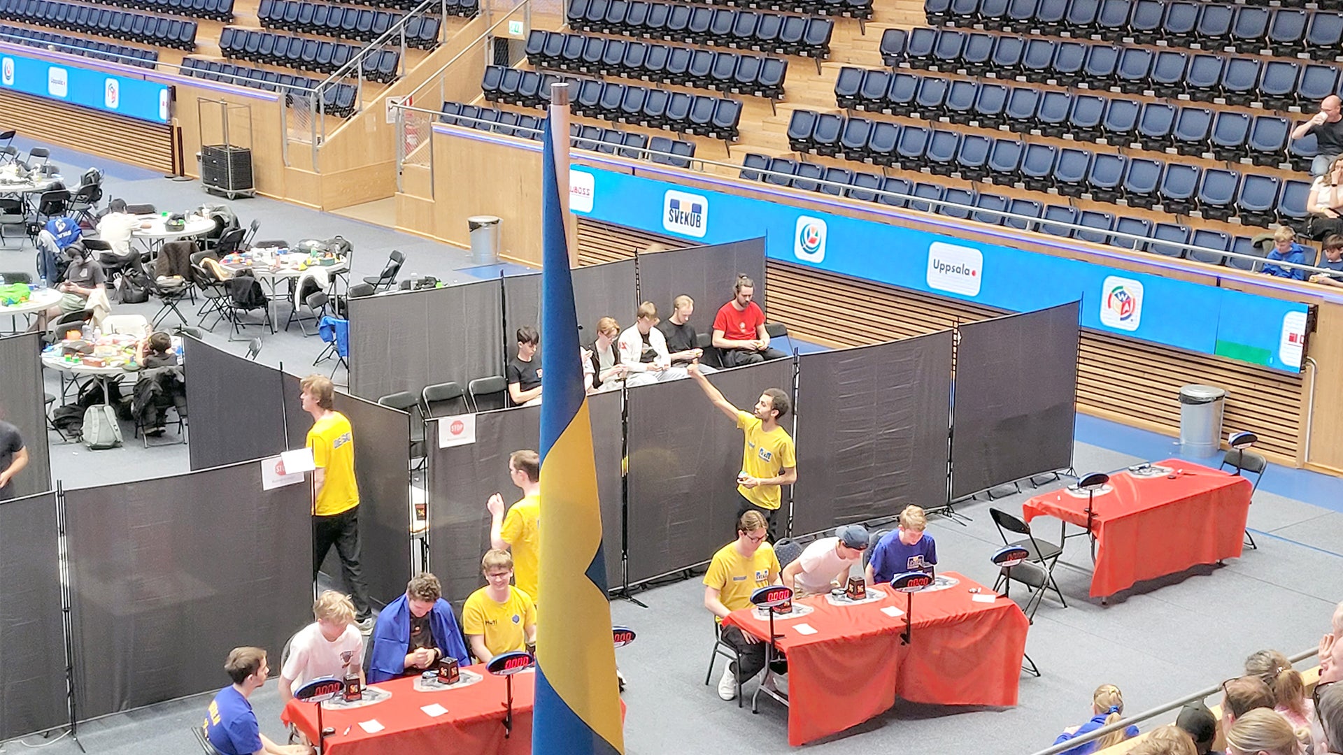 Utställningsbås på ett evenemang i en idrottshall med besökare och personal i gula västar vid röda bord.