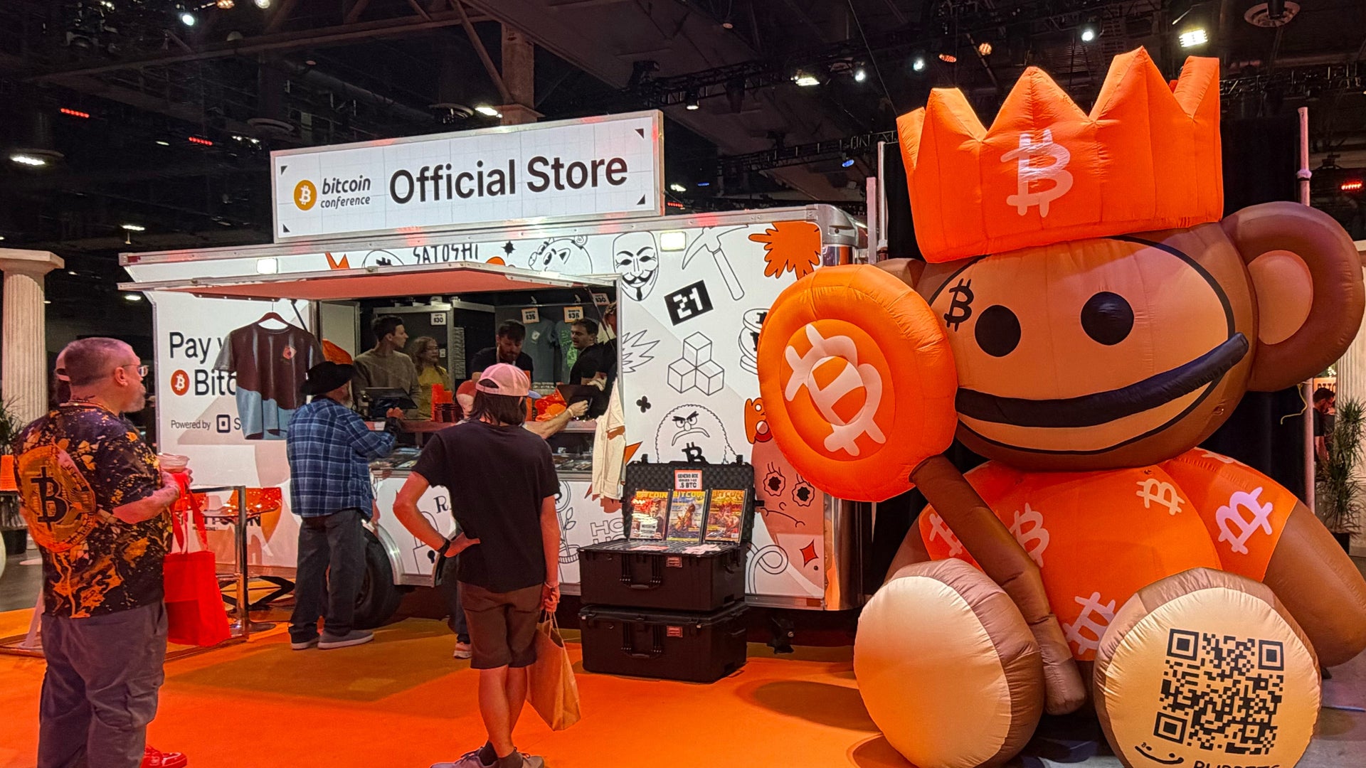 En stor orange Bitcoin-maskot med leende ansikte står vid en utställningsmonter märkt 'Official Store' på en mässa. Människor besöker standen runt omkring.