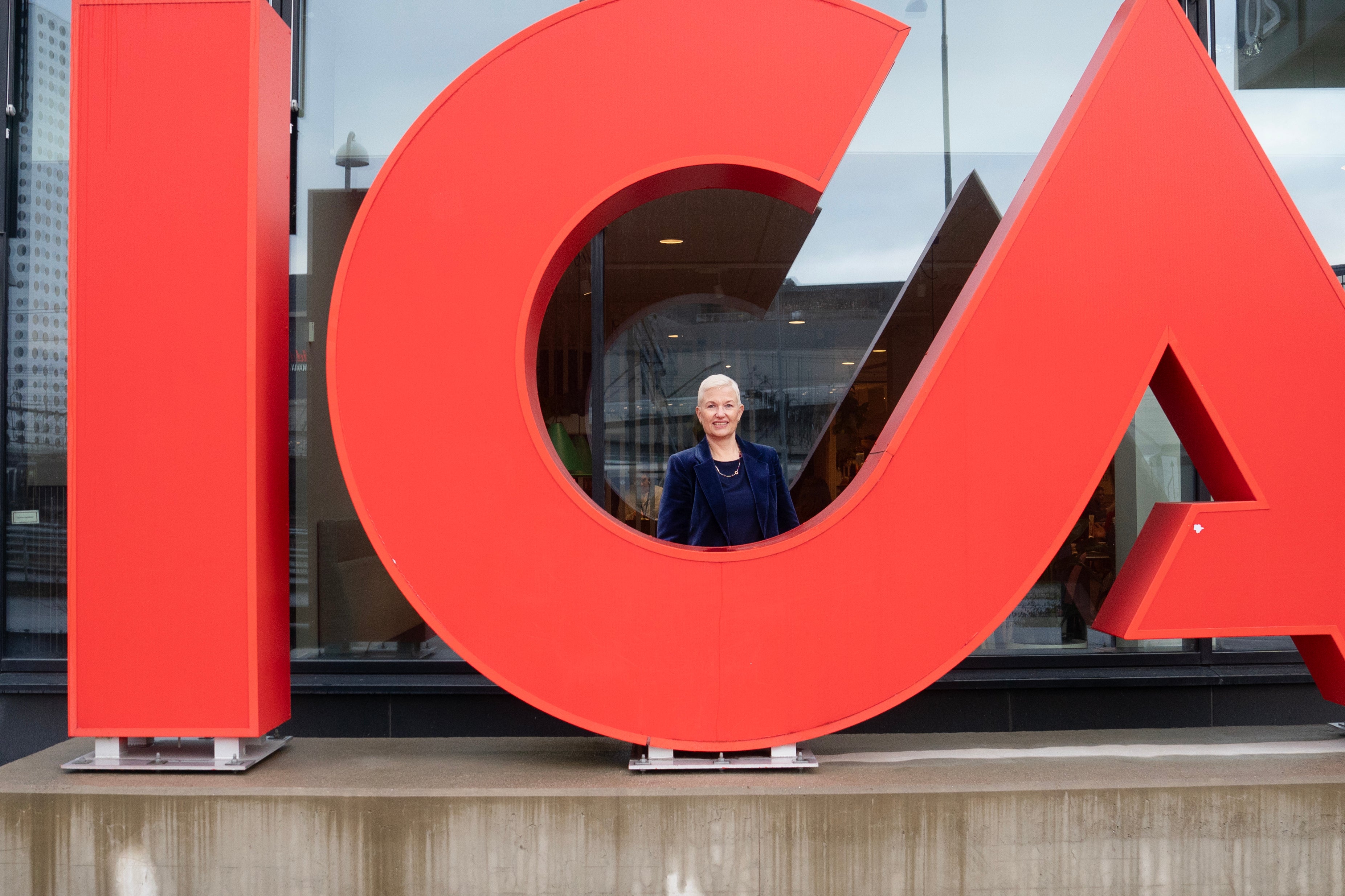 En stor, röd staty över Icas logotyp framför ett fönster i Arenastaden. Bakom bokstaven C står Icas koncernchef Nina Jönsson, en blond kvinna.