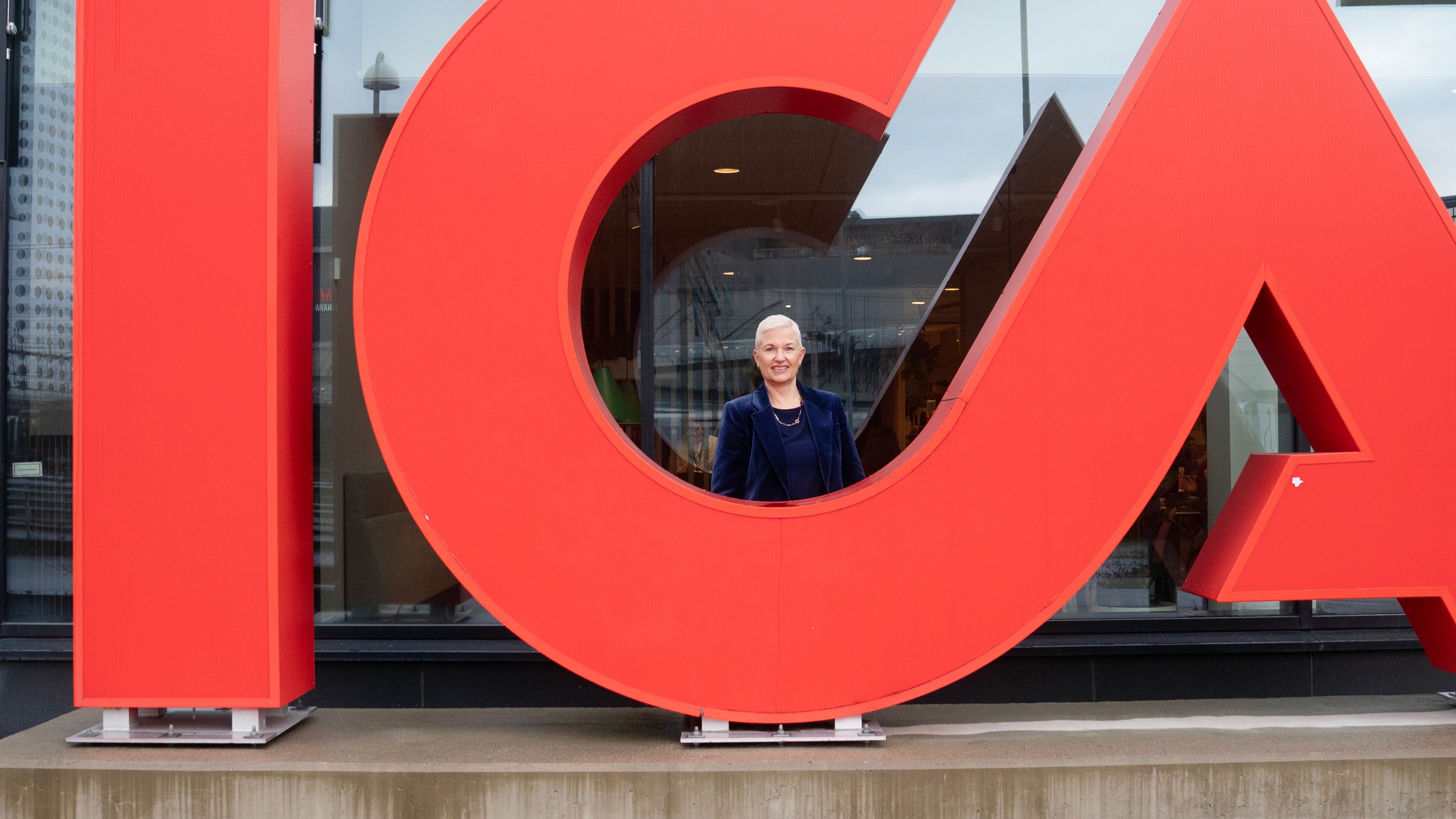 En stor, röd staty över Icas logotyp framför ett fönster i Arenastaden. Bakom bokstaven C står Icas koncernchef Nina Jönsson, en blond kvinna.