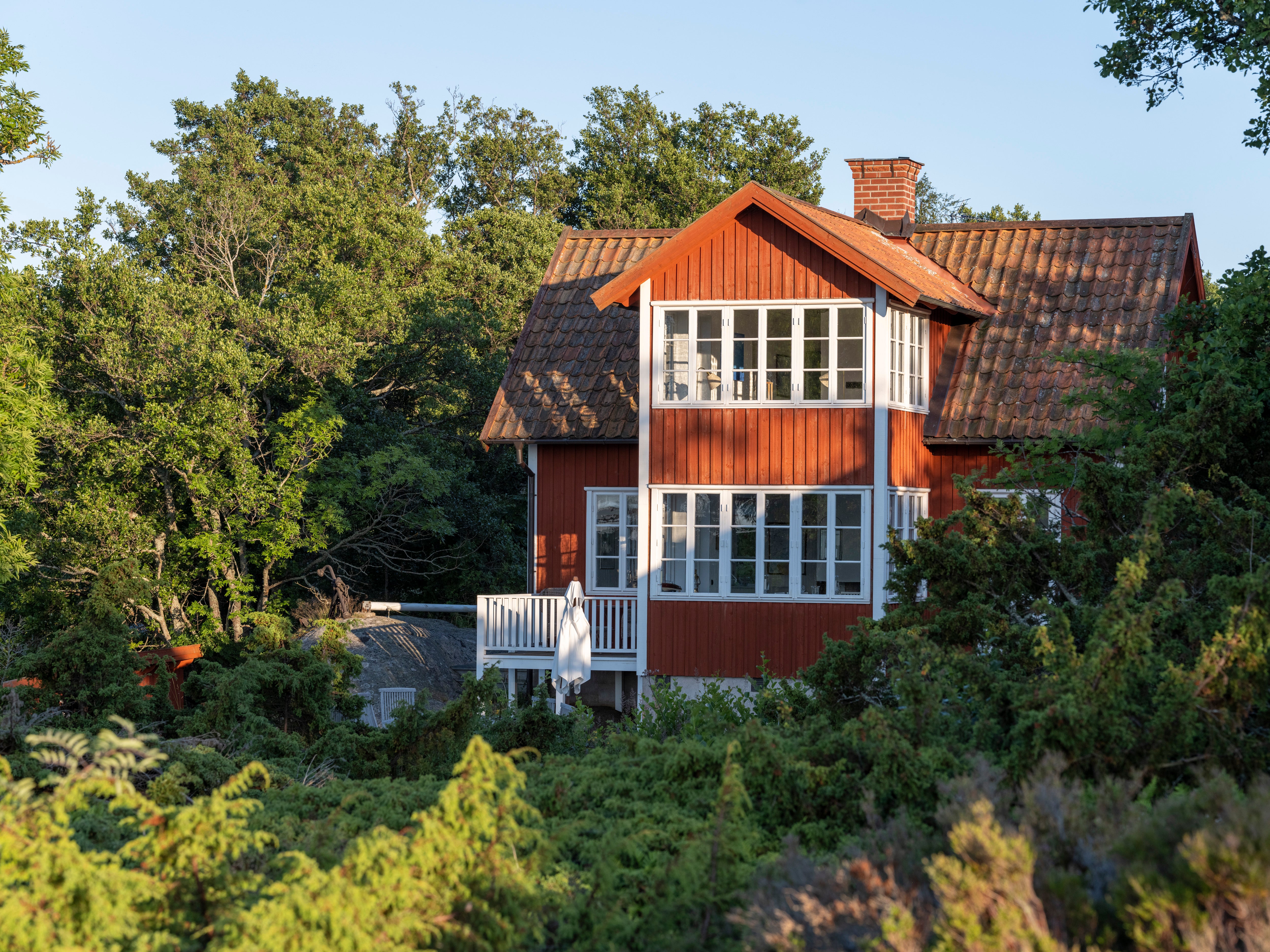 Rött trähus med vita fönster och vita detaljer, omgivet av träd och grönska.
