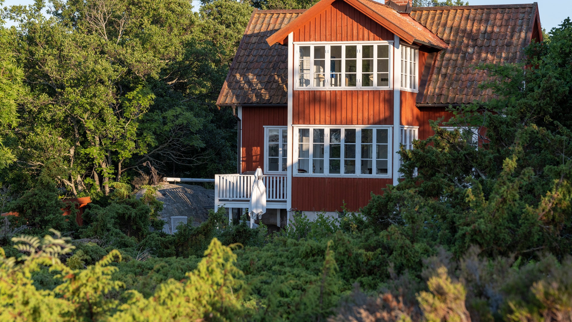 Rött trähus med vita fönster och vita detaljer, omgivet av träd och grönska.