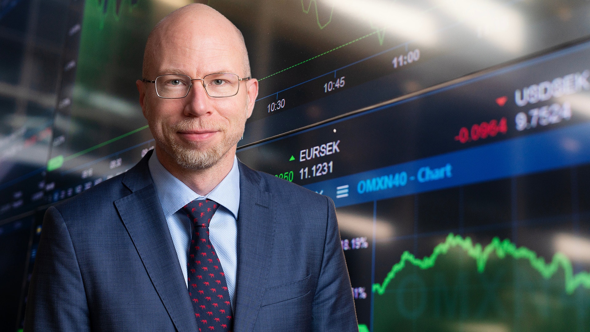 Porträtt av en man i blå kostym och glasögon framför en finansiell handelsskärm med aktiekurser och diagram.