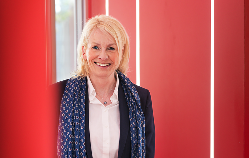 Bodil Sonesson står i ett rum med röda väggar och ler in i kameran.