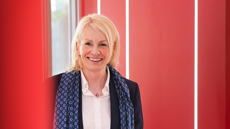 Bodil Sonesson står i ett rum med röda väggar och ler in i kameran.